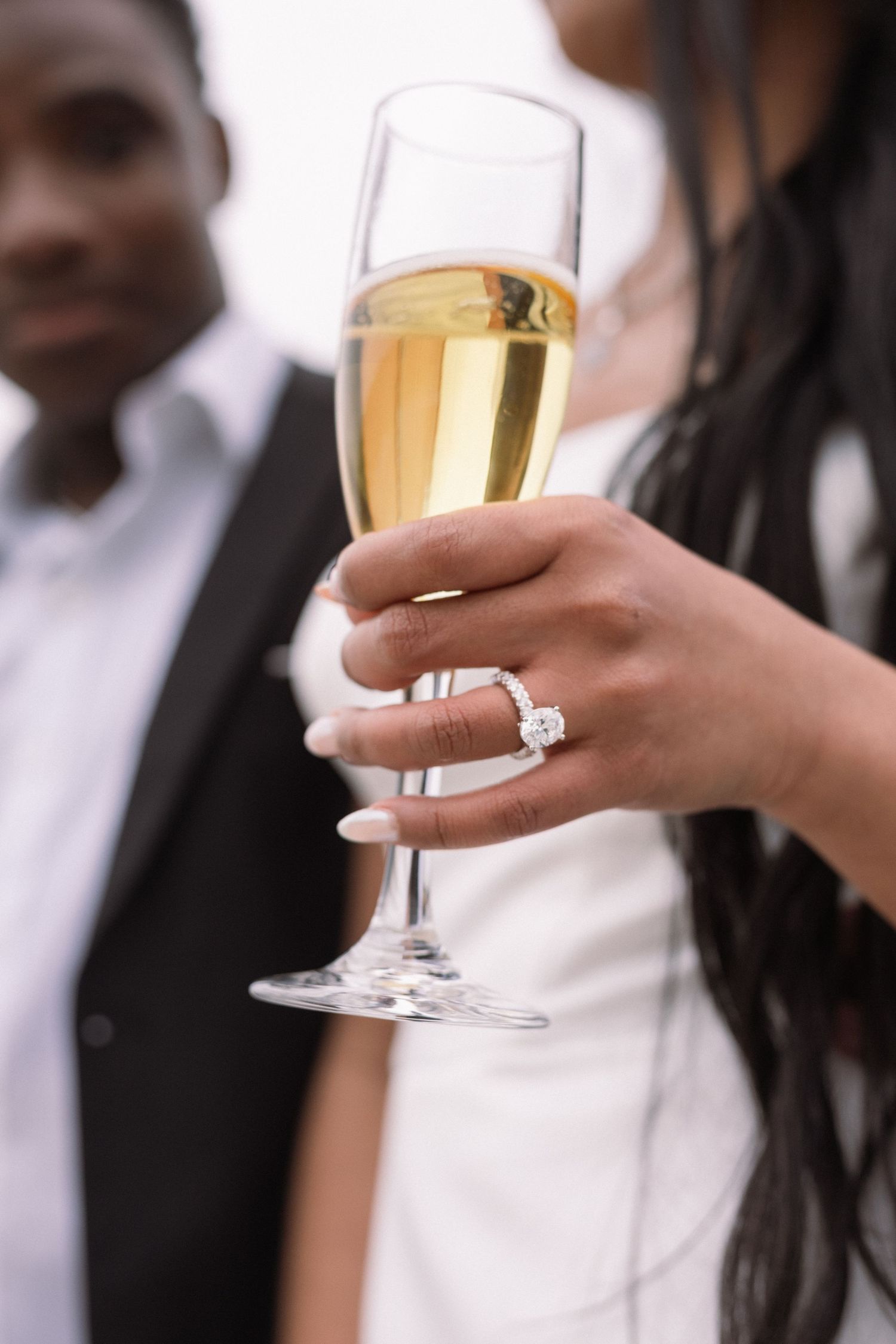 Close-up of a woman's hand holding a champagne flute, showcasing a diamond engagement ring, with a couple in the background.
