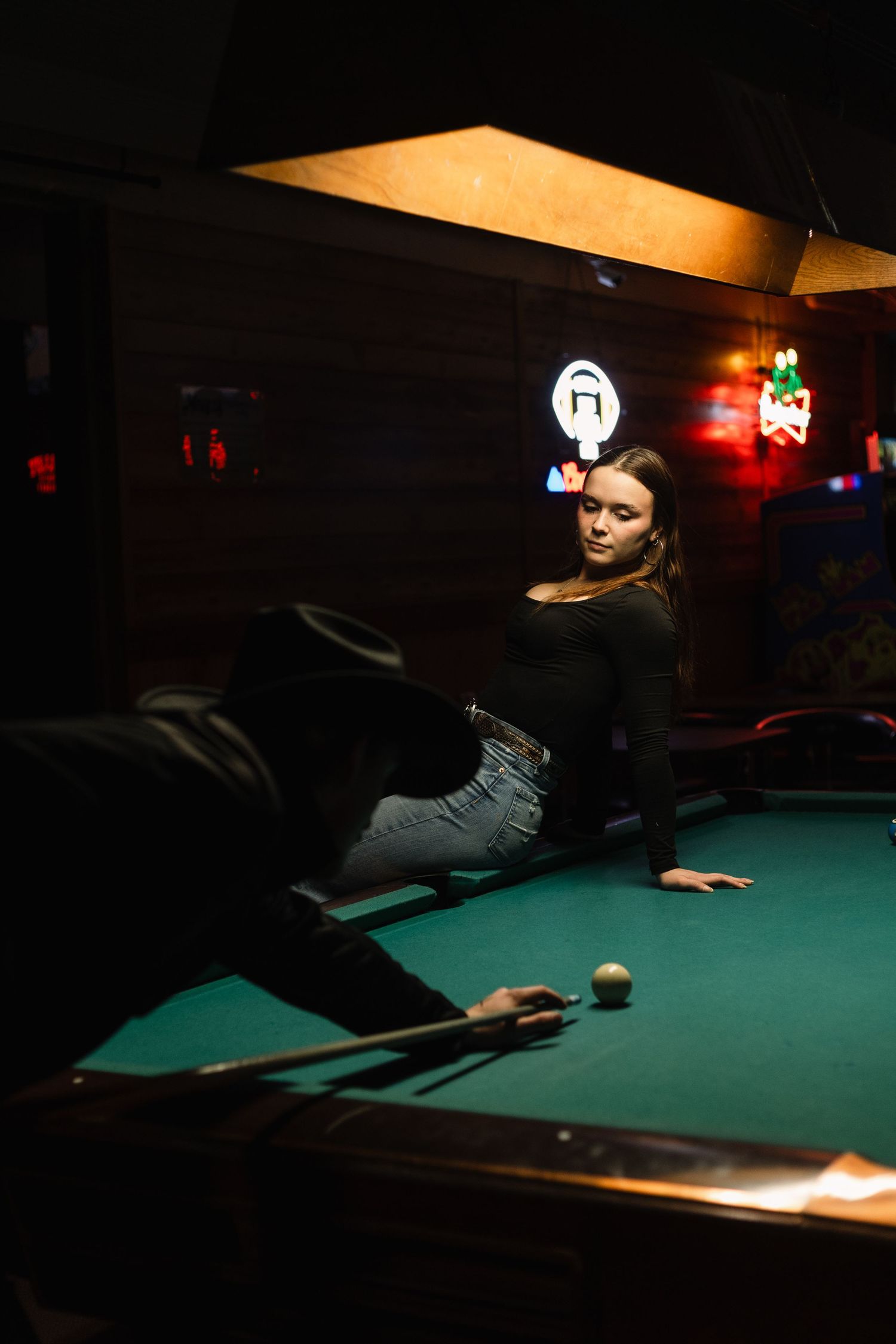 A person plays pool in a dimly lit bar with dramatic shadows cast across the green felt table.