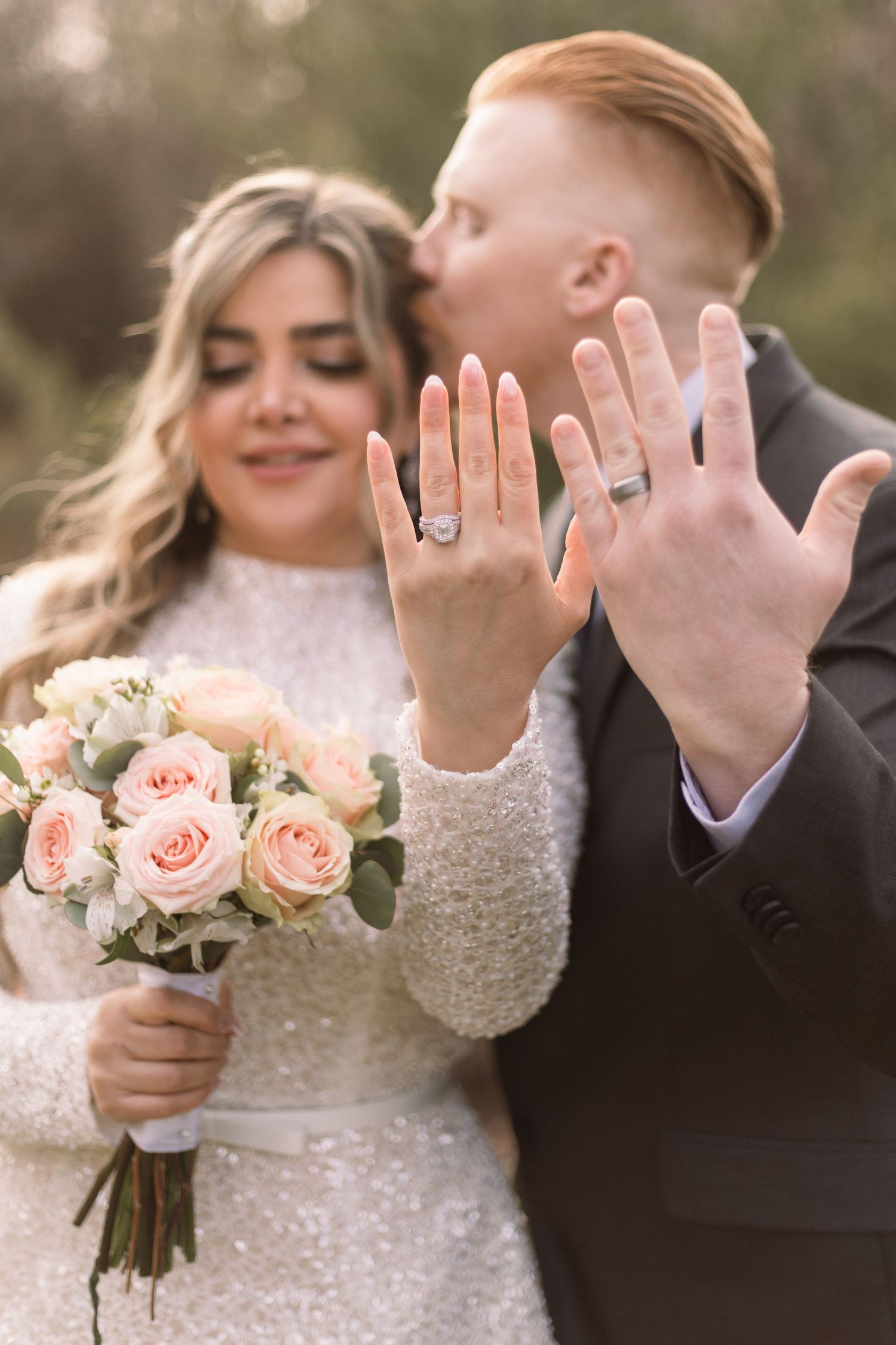 Newlywed couple shows off their wedding rings while holding a pink rose bouquet in an outdoor setting.
