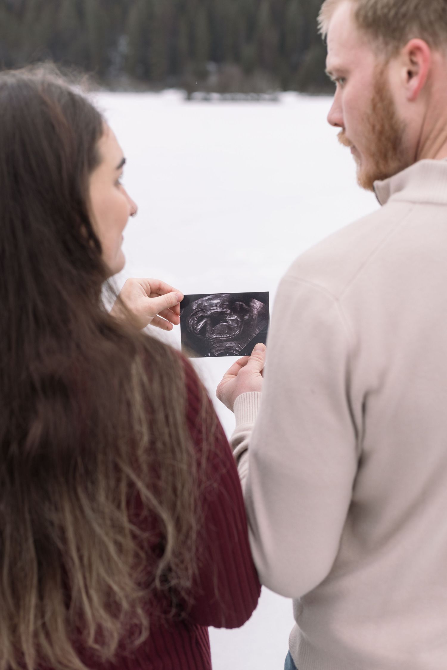 Couple looking at ultrasound photos during winter maternity shoot in snowy landscape.
