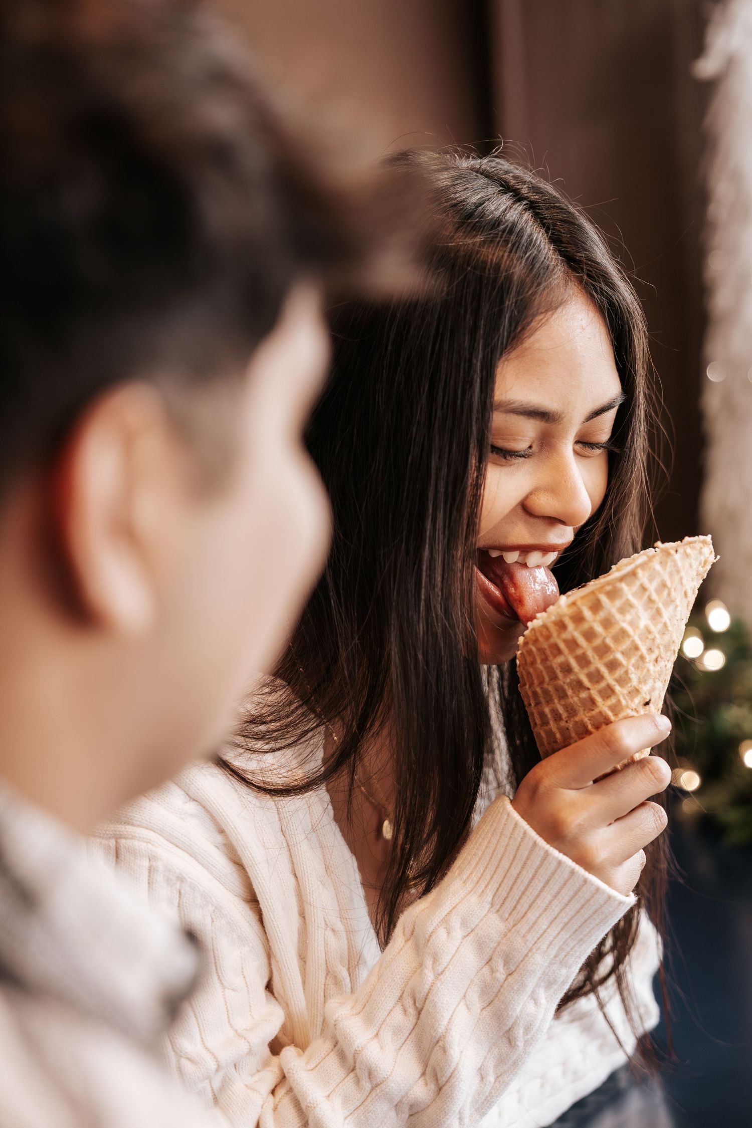 A woman in a white sweater enjoys eating a waffle cone ice cream with blurred bokeh lights in the background.