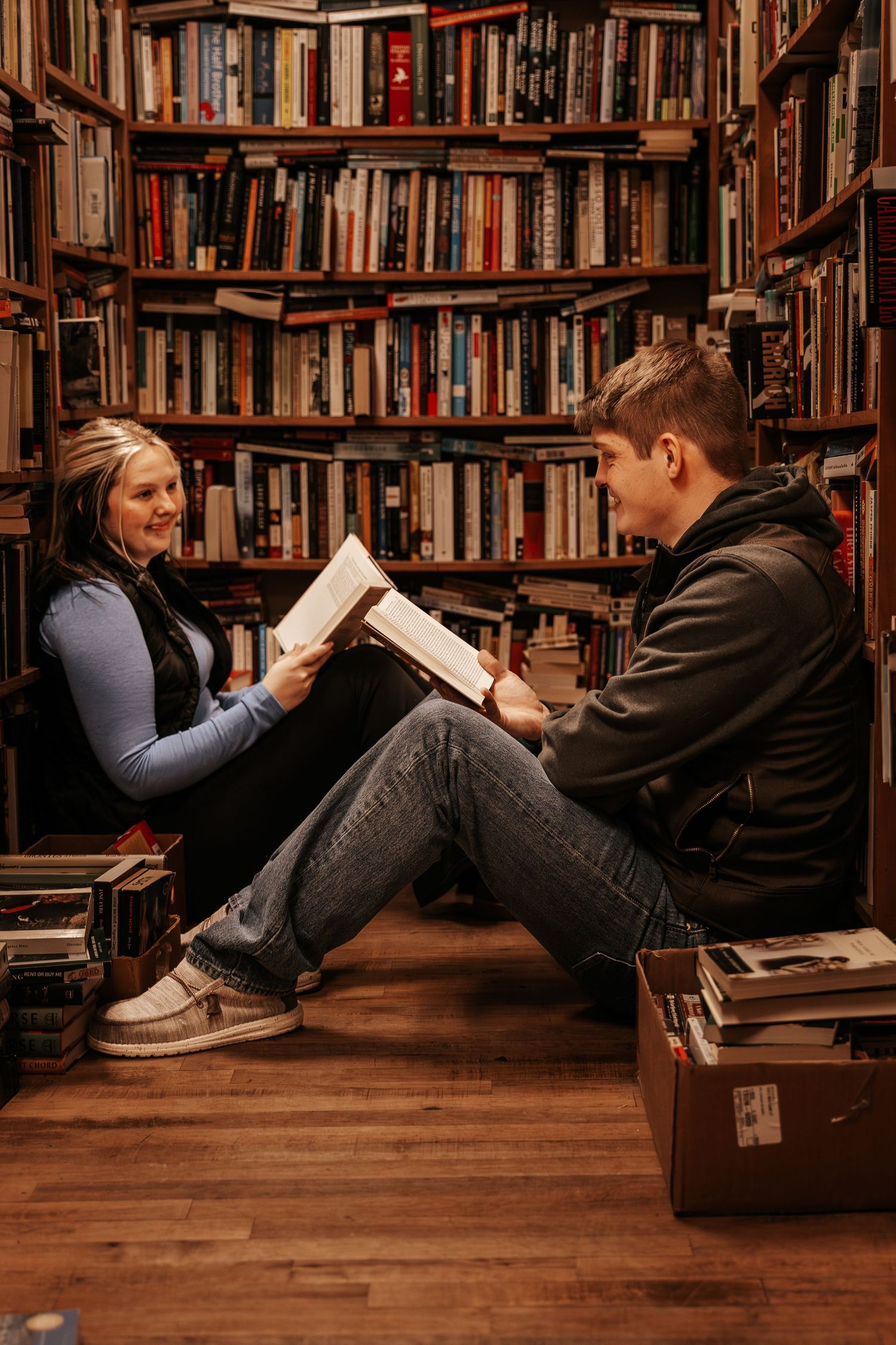 Two people reading books while sitting on wooden floor in cozy library with floor-to-ceiling bookshelves.