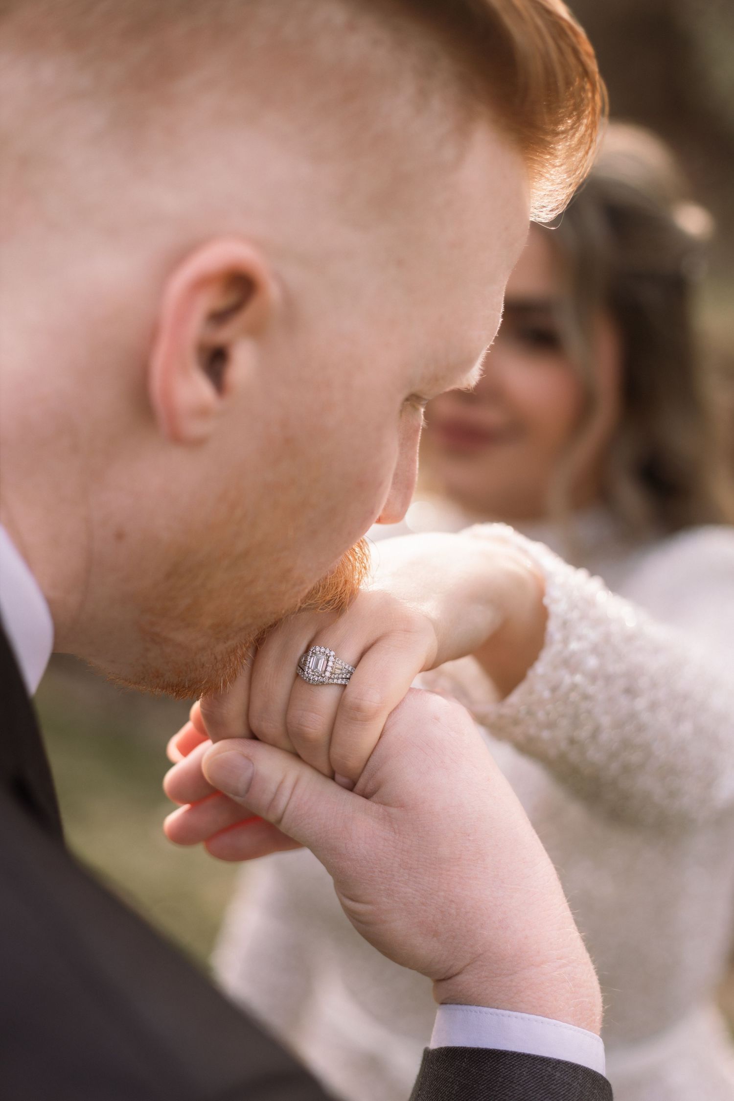 Groom with red hair kisses bride's hand adorned with a diamond engagement ring during romantic wedding moment.
