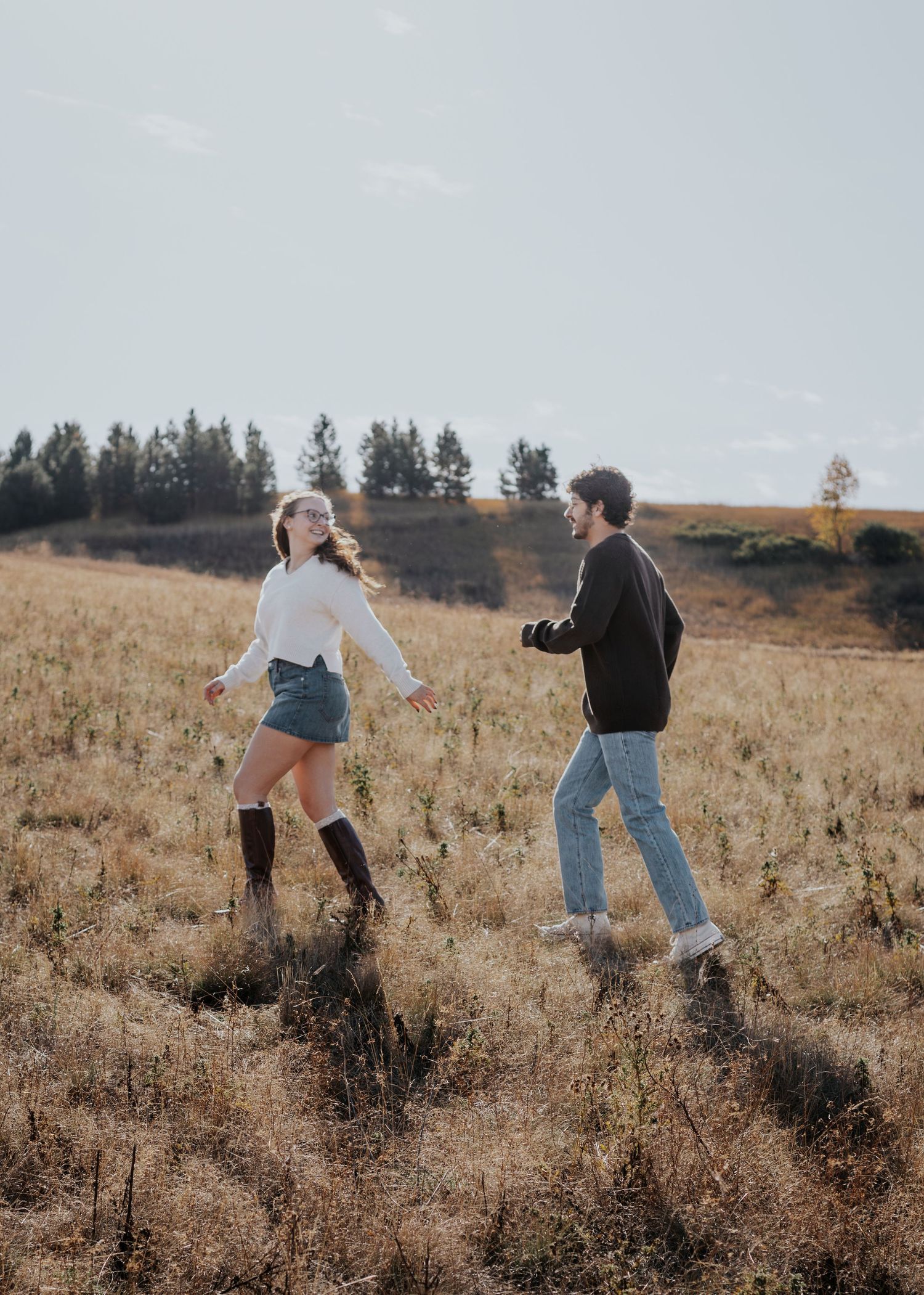 Two people dancing and having fun in a golden field with evergreen trees on the hillside during sunset.