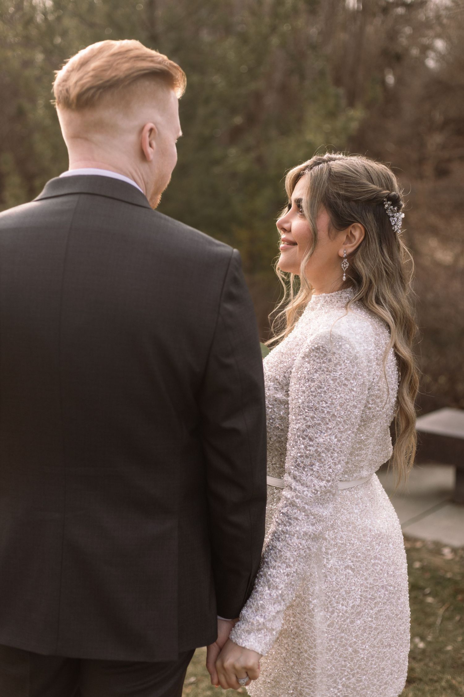 A couple in formal wedding attire holds hands while walking together in an outdoor setting during autumn.