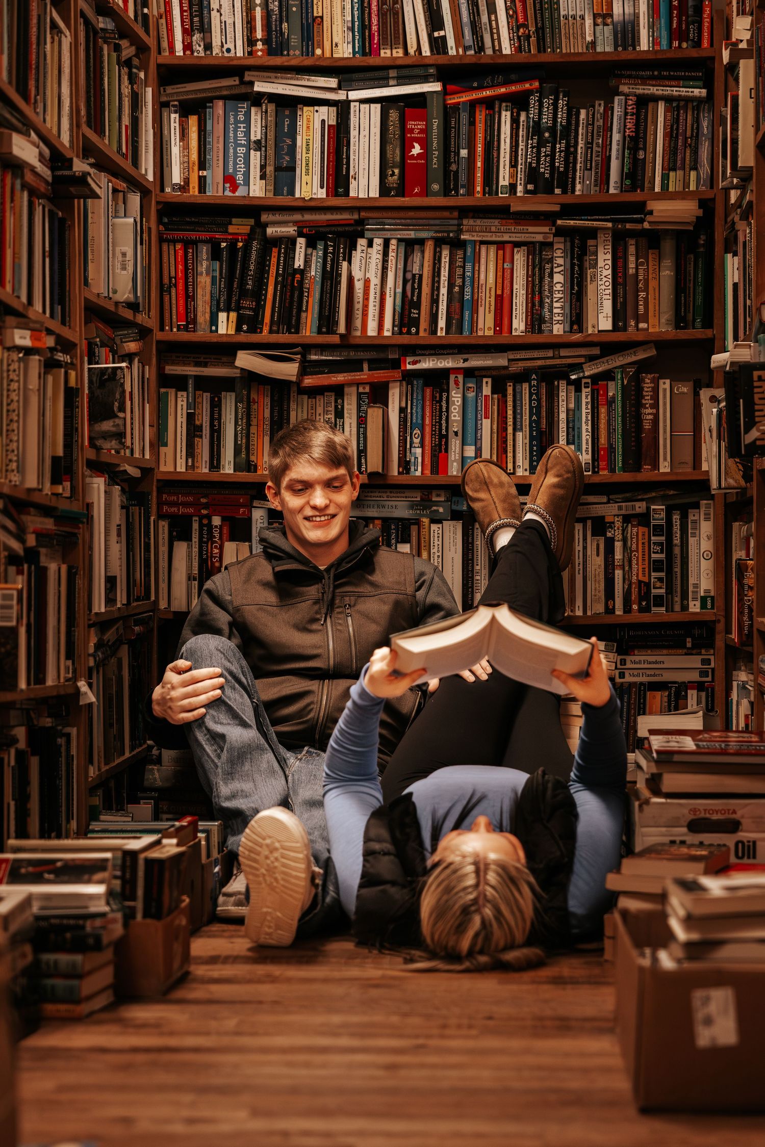 A person sits comfortably on a wooden floor surrounded by towering bookshelves in a cozy library setting.