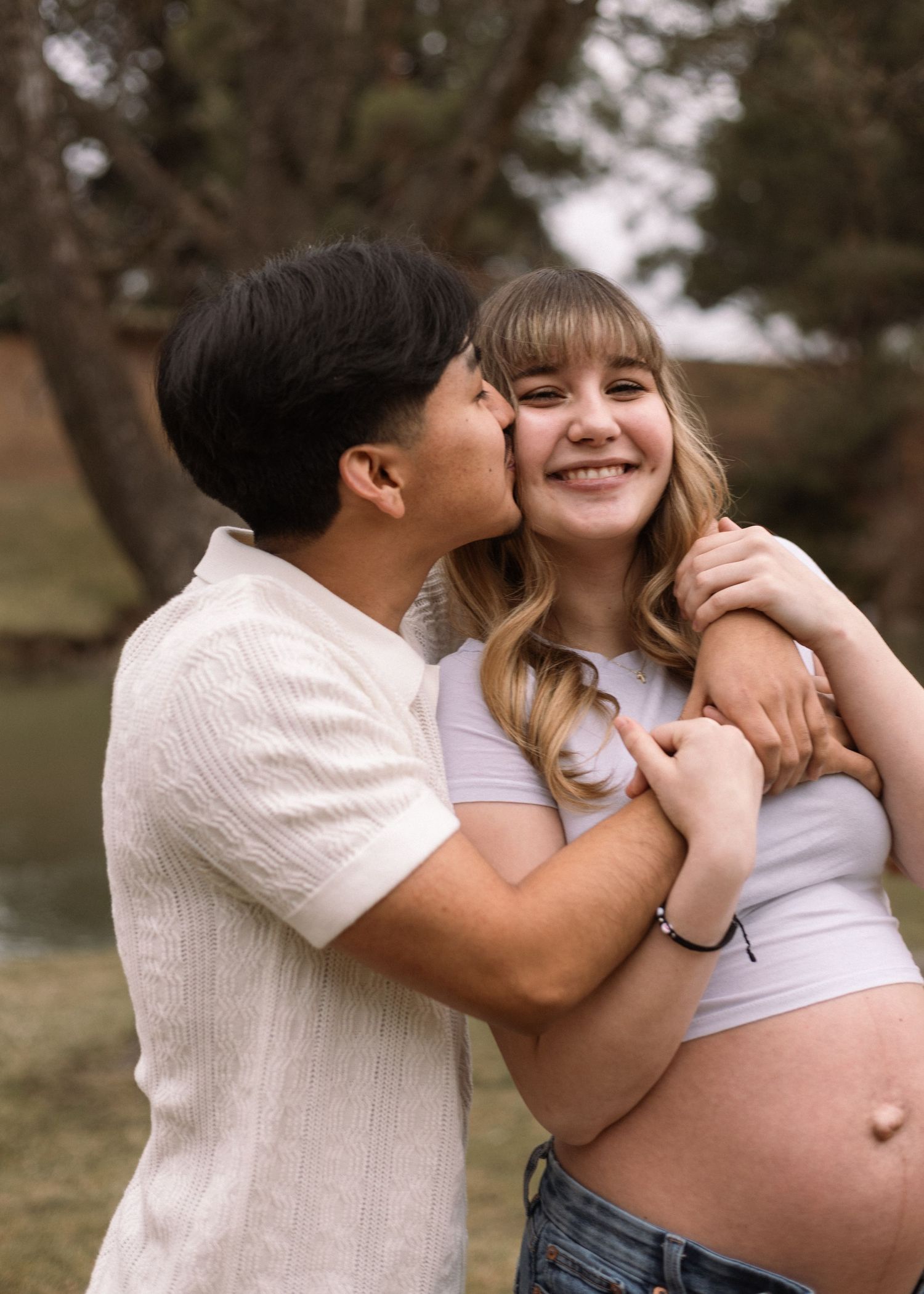 A loving couple embraces outdoors while showing off a pregnant belly in casual attire.