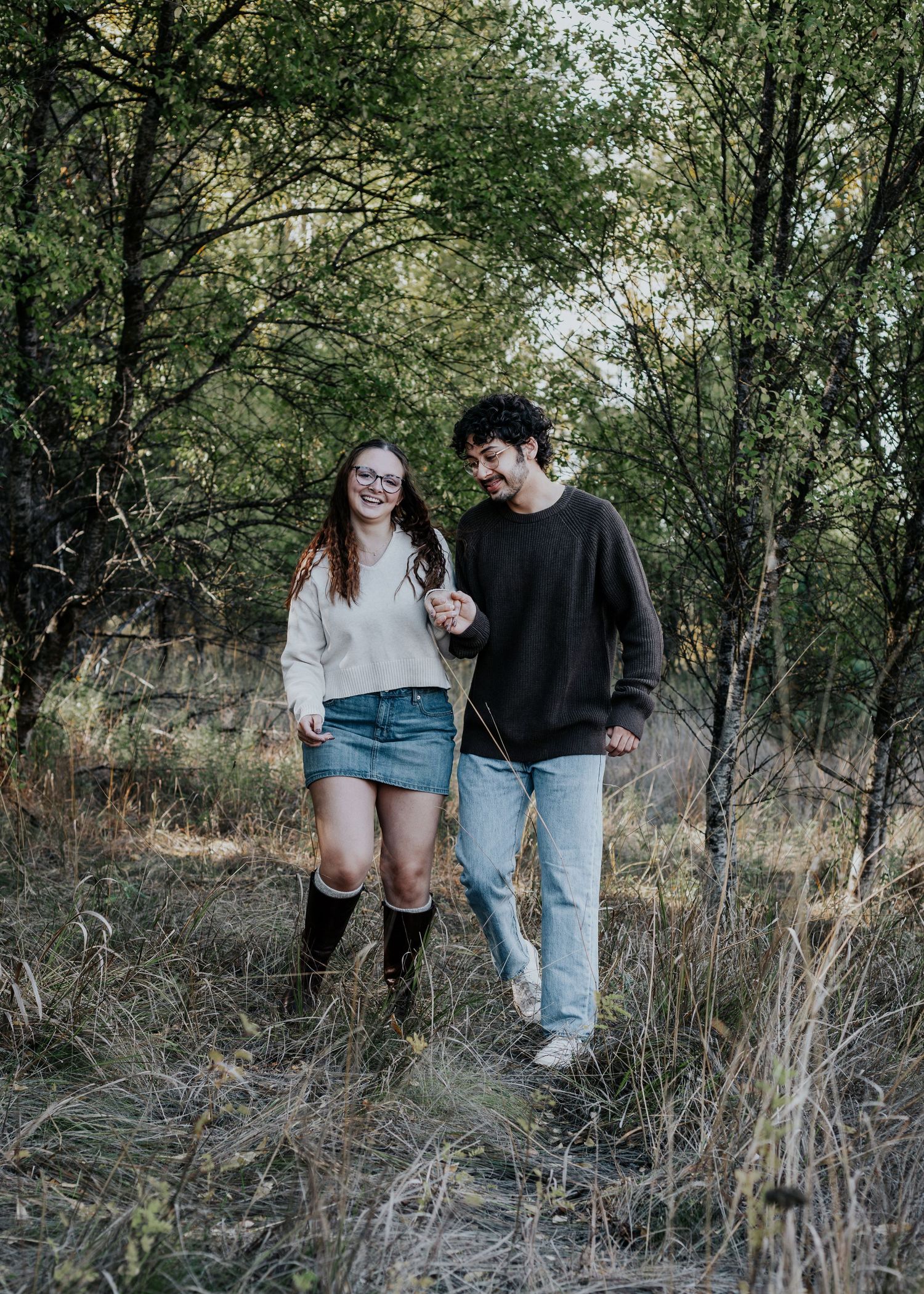 A couple walks together through tall grass and trees on an autumn day wearing casual outfits.