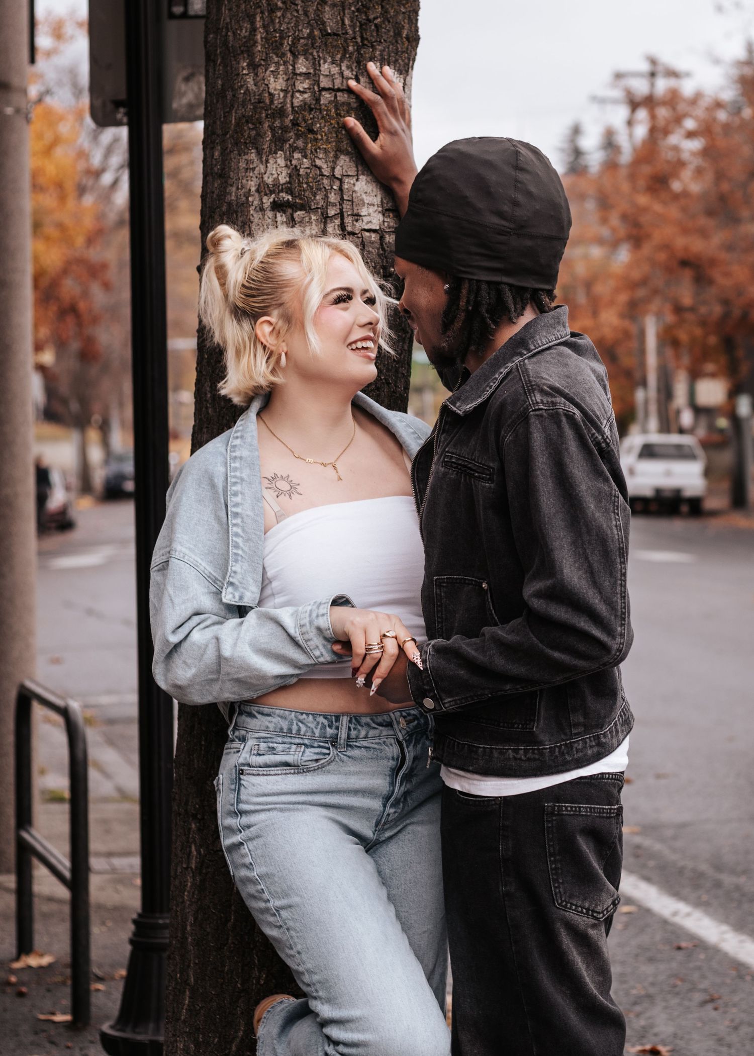 A couple in denim and black clothing embraces against a tree trunk on an autumn street with orange foliage.