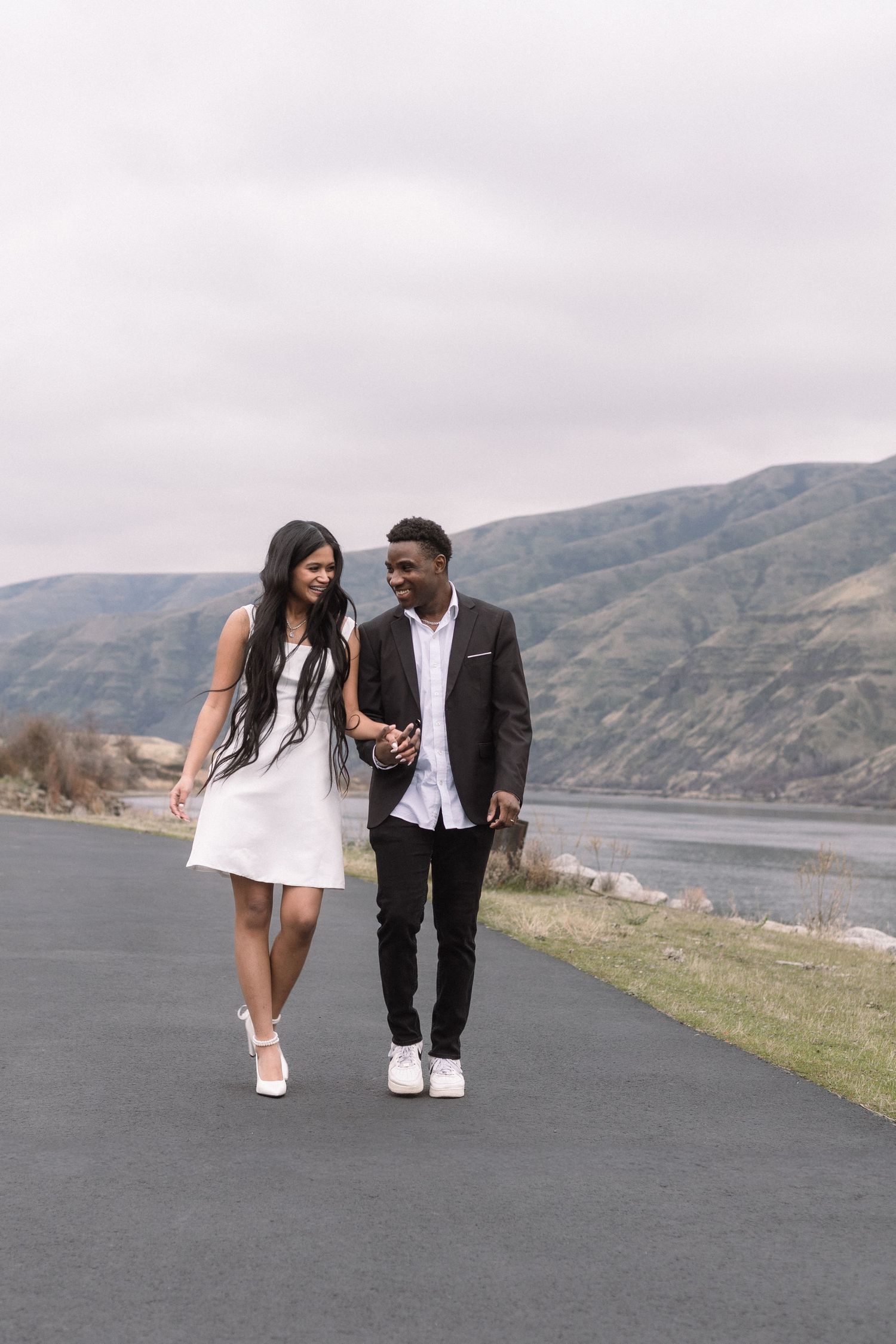 A happy interracial couple walks hand-in-hand along a scenic mountain lakeside path under an overcast sky.