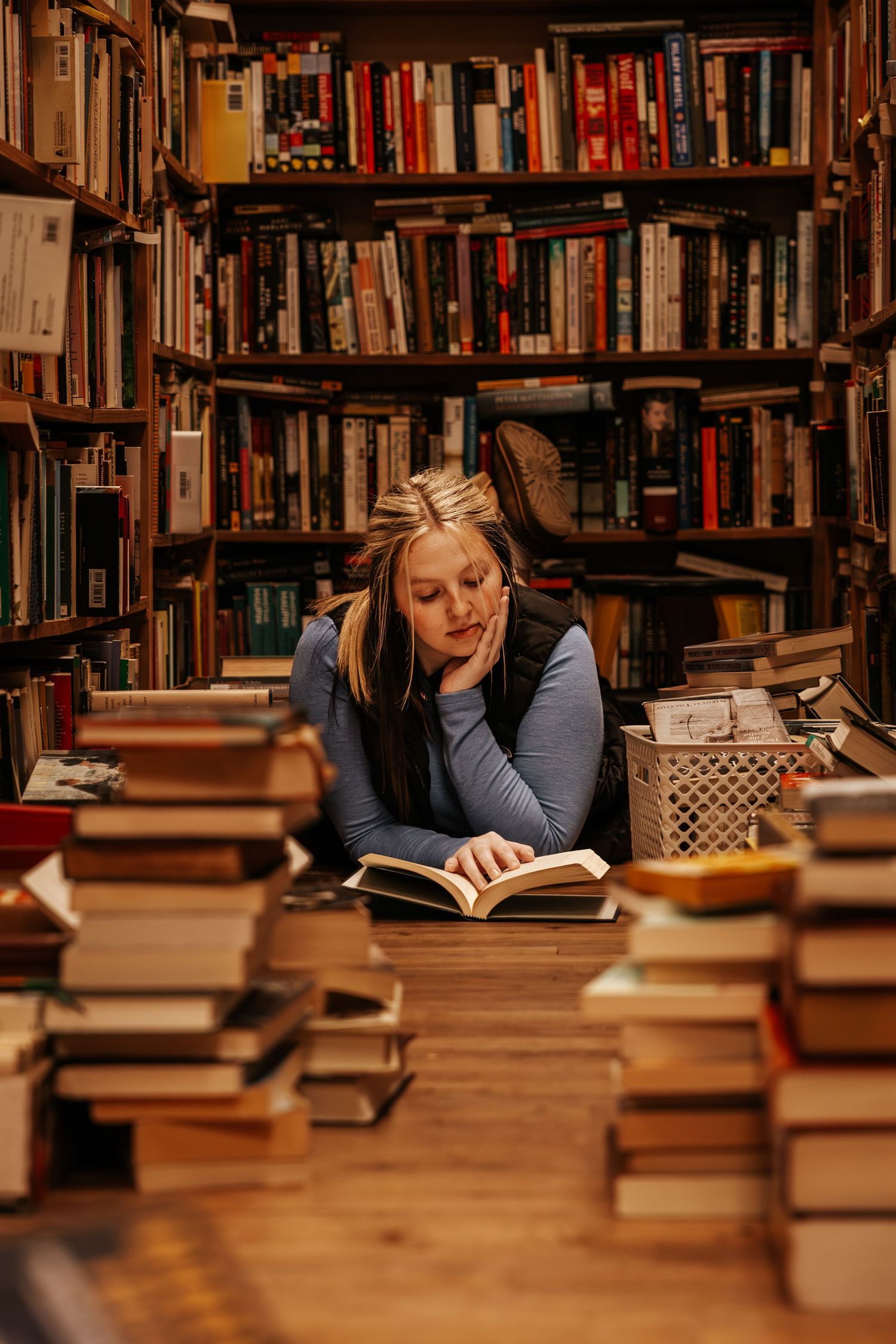 Student studying late at night in library surrounded by stacks of books on wooden table.