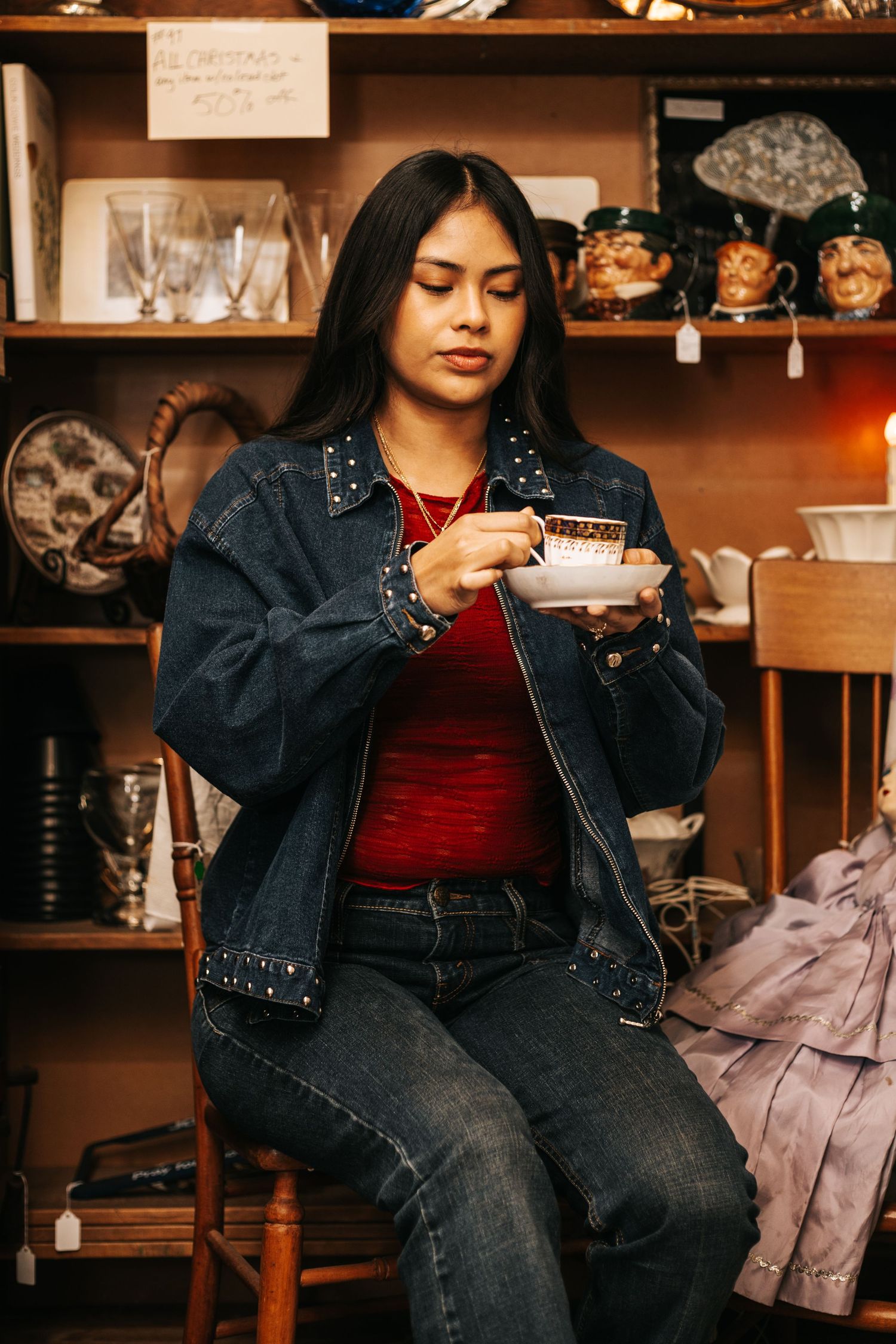 Person in denim jacket and red top sits in vintage shop surrounded by glassware and collectibles.