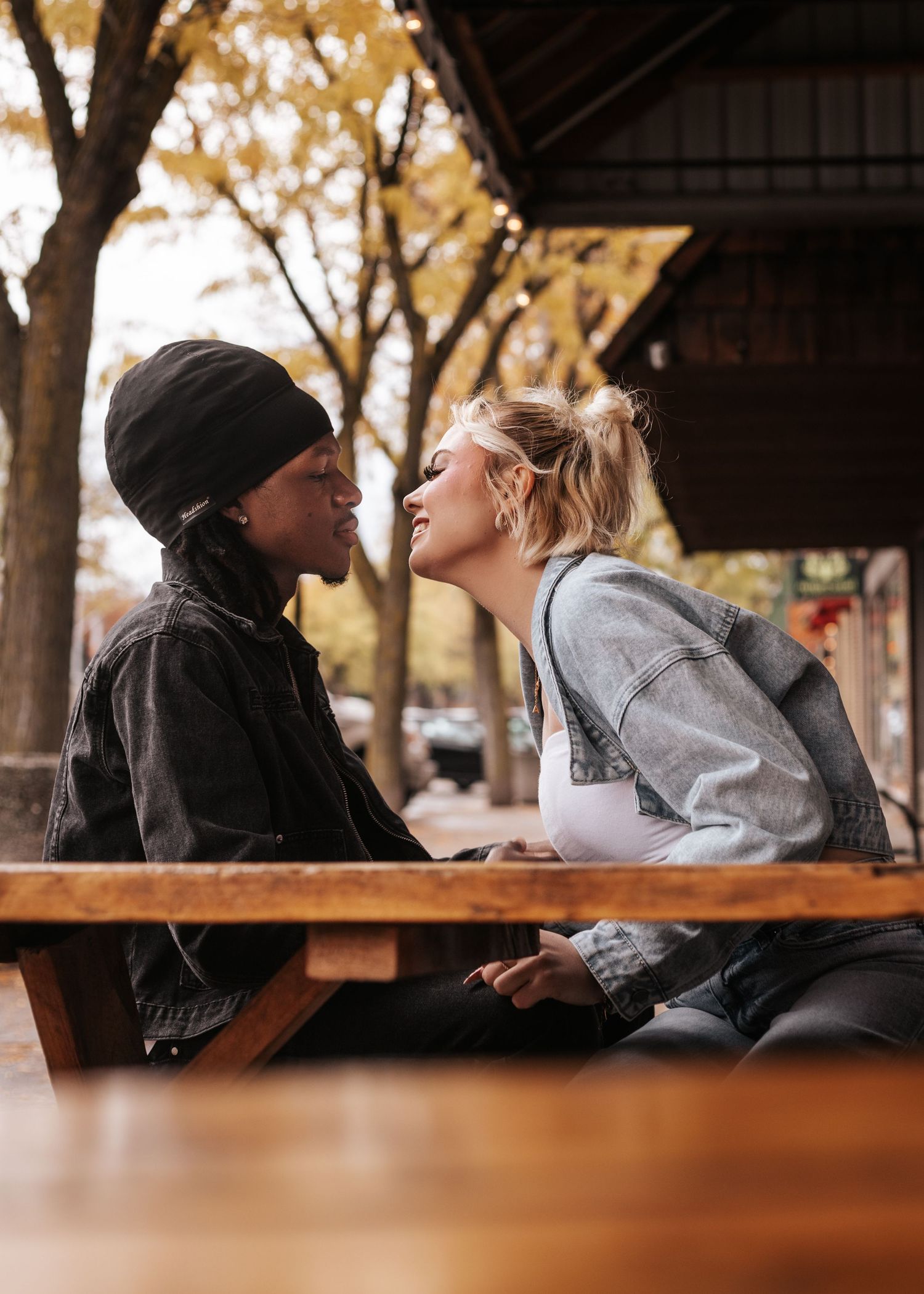 Two people in winter hats share a tender moment while sitting at a wooden picnic table on an autumn day.