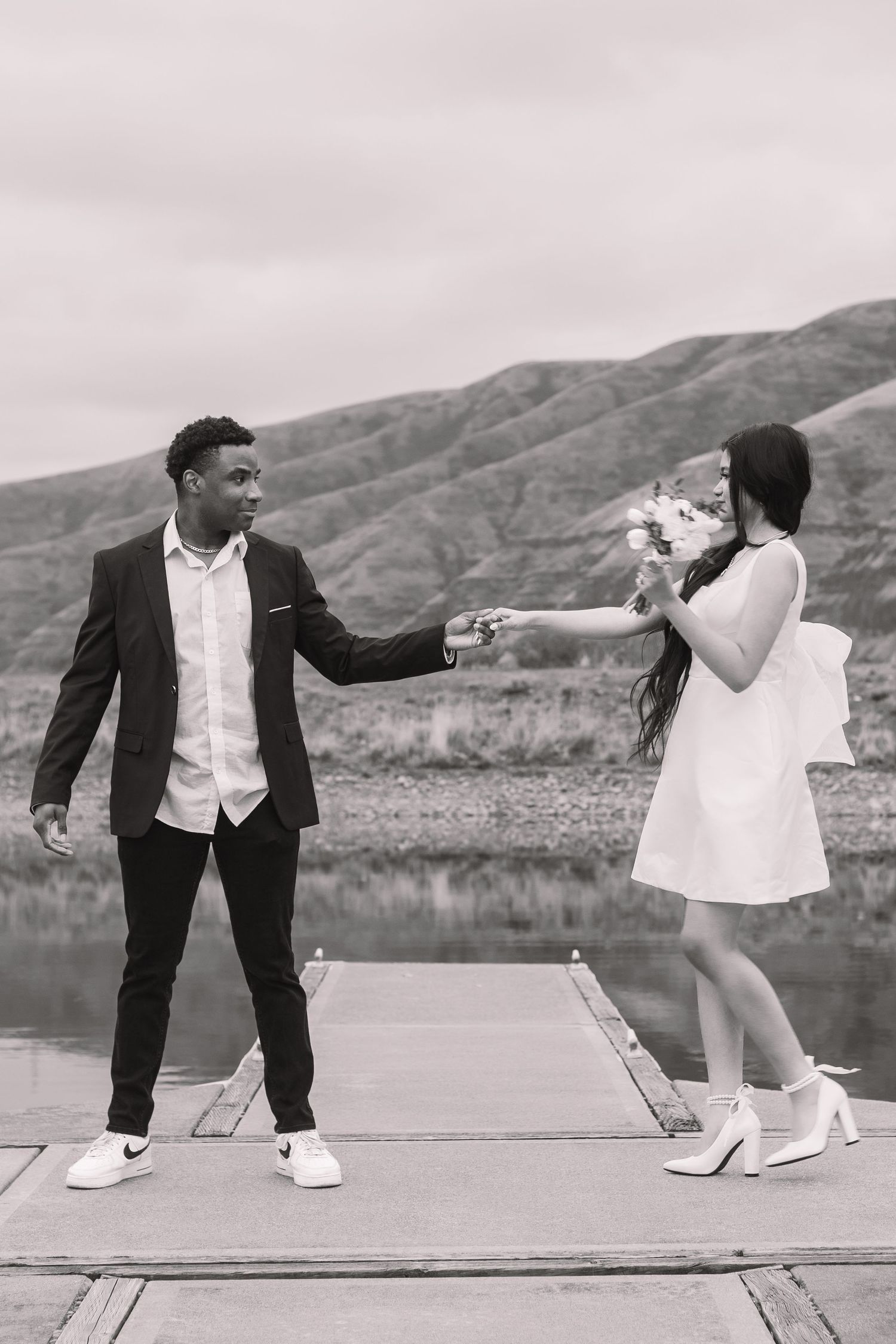 A playful couple in wedding attire dances on a dock by a mountain lake.