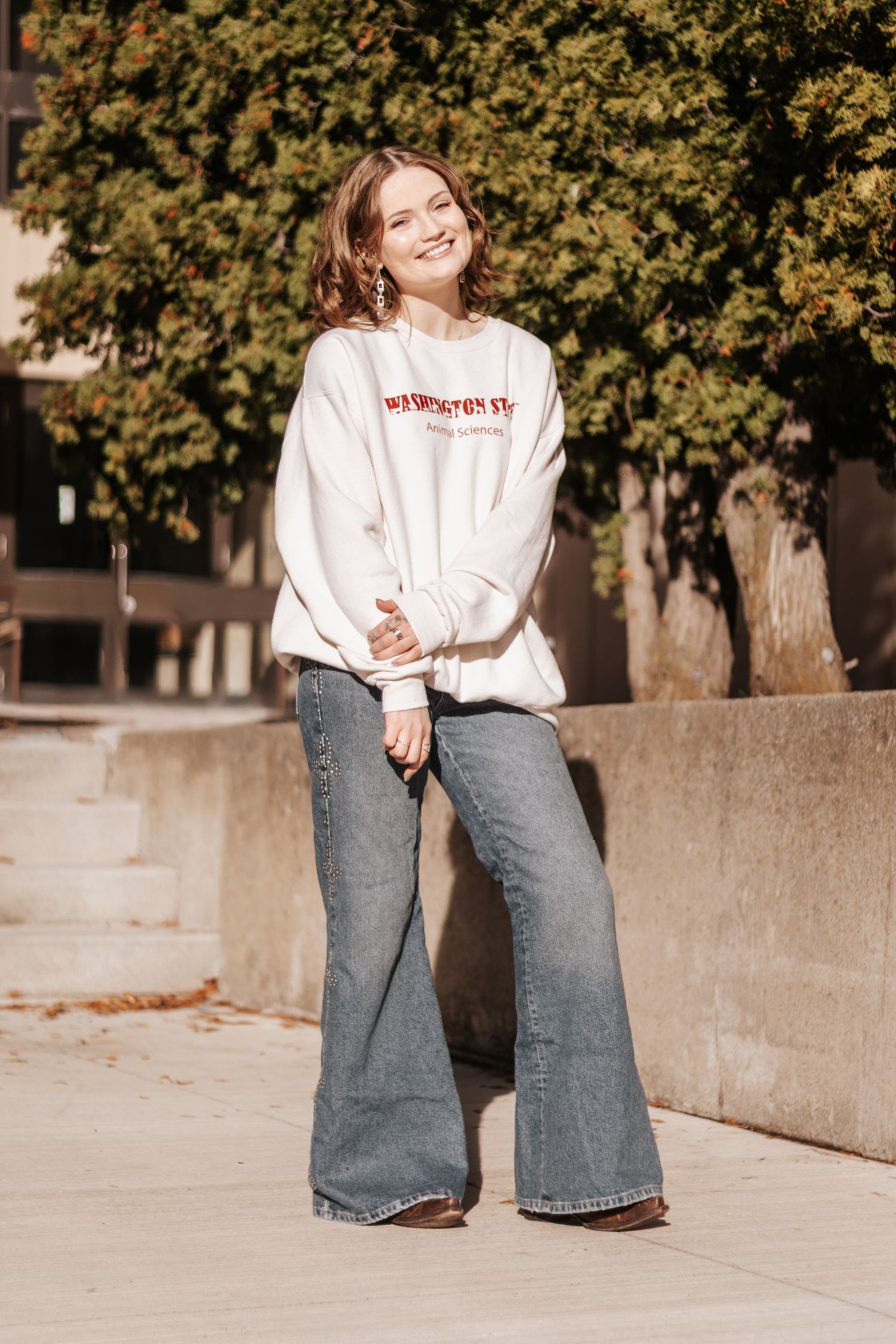 A person in a white sweatshirt and flared jeans poses casually on a concrete ledge with autumn trees in the background.