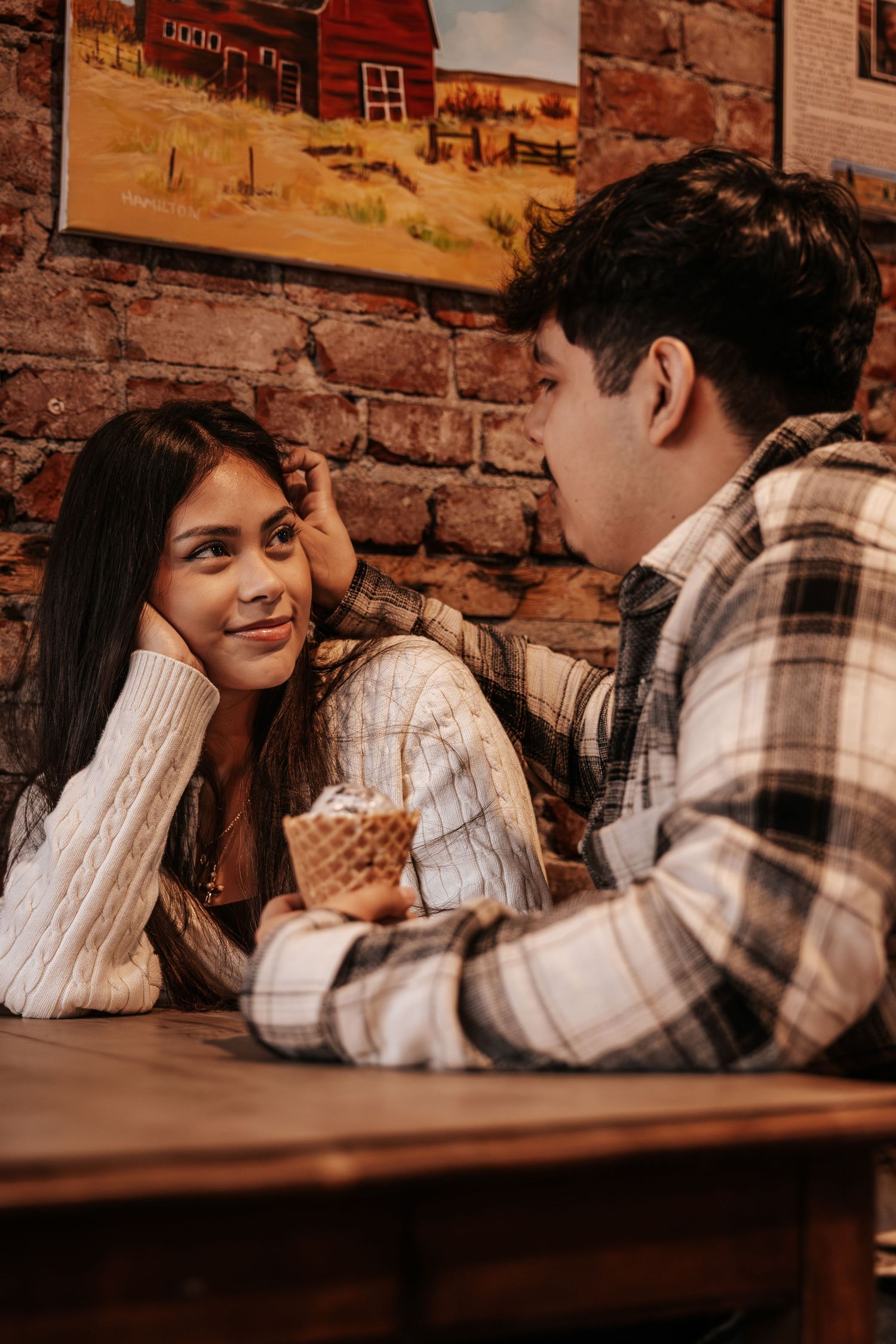 A romantic couple sits at a rustic wooden table against a brick wall, sharing intimate moments in warm lighting.
