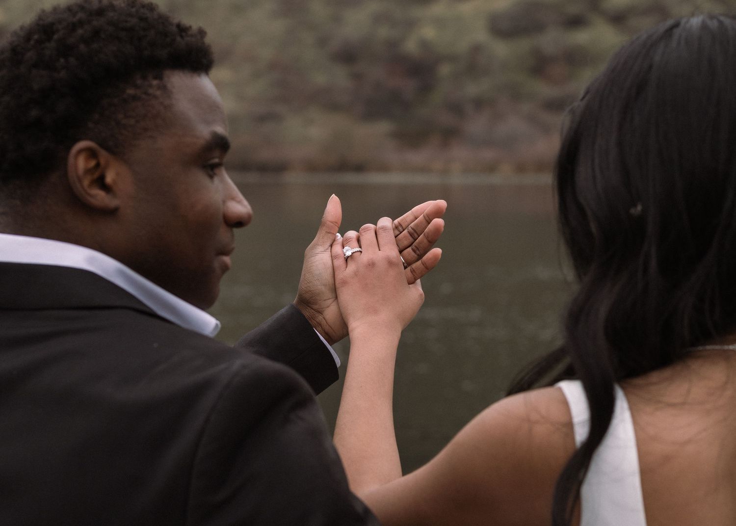 A couple shares a romantic moment examining an engagement ring outdoors near a lake.