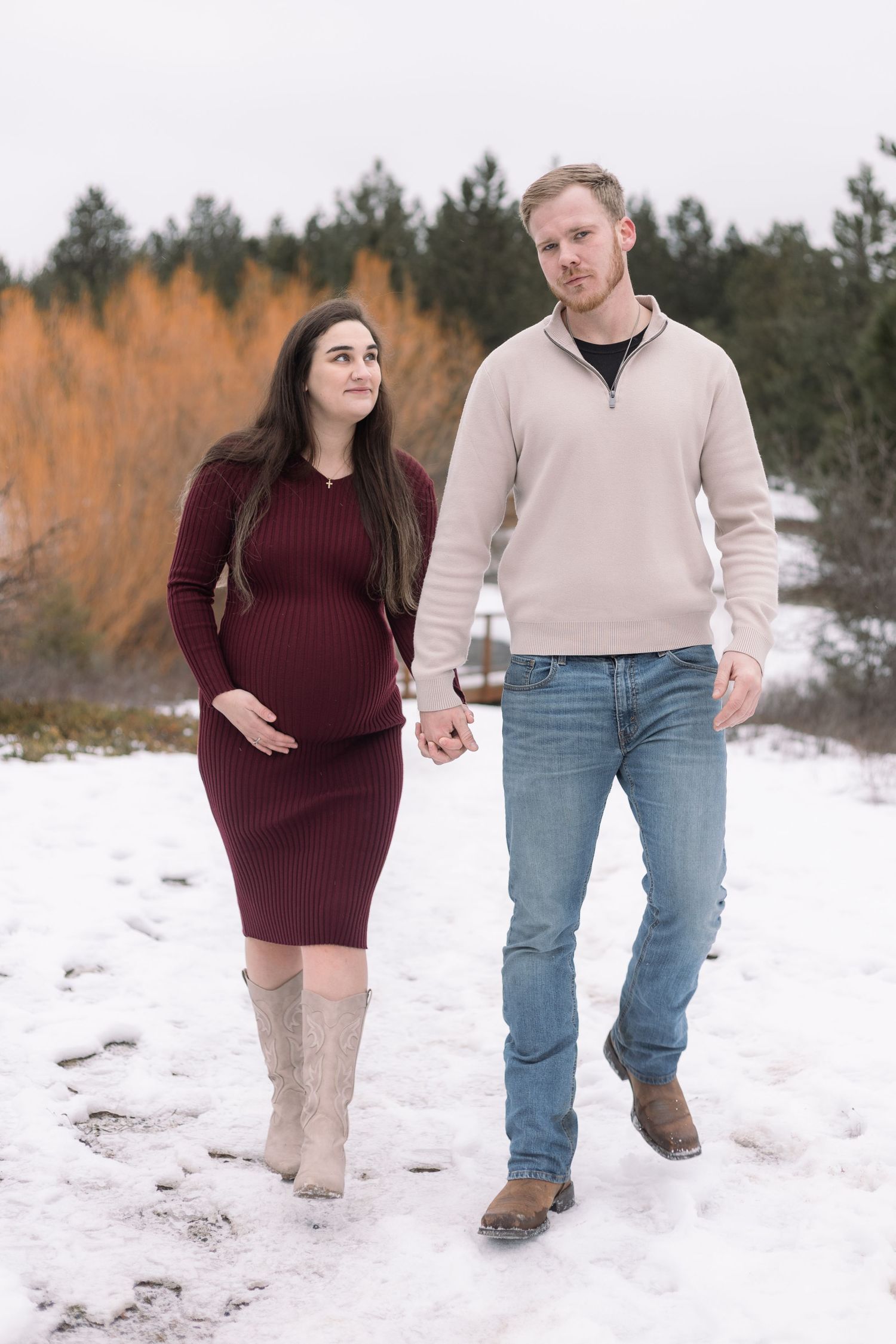 Couple walking hand in hand through snow during winter maternity photoshoot.