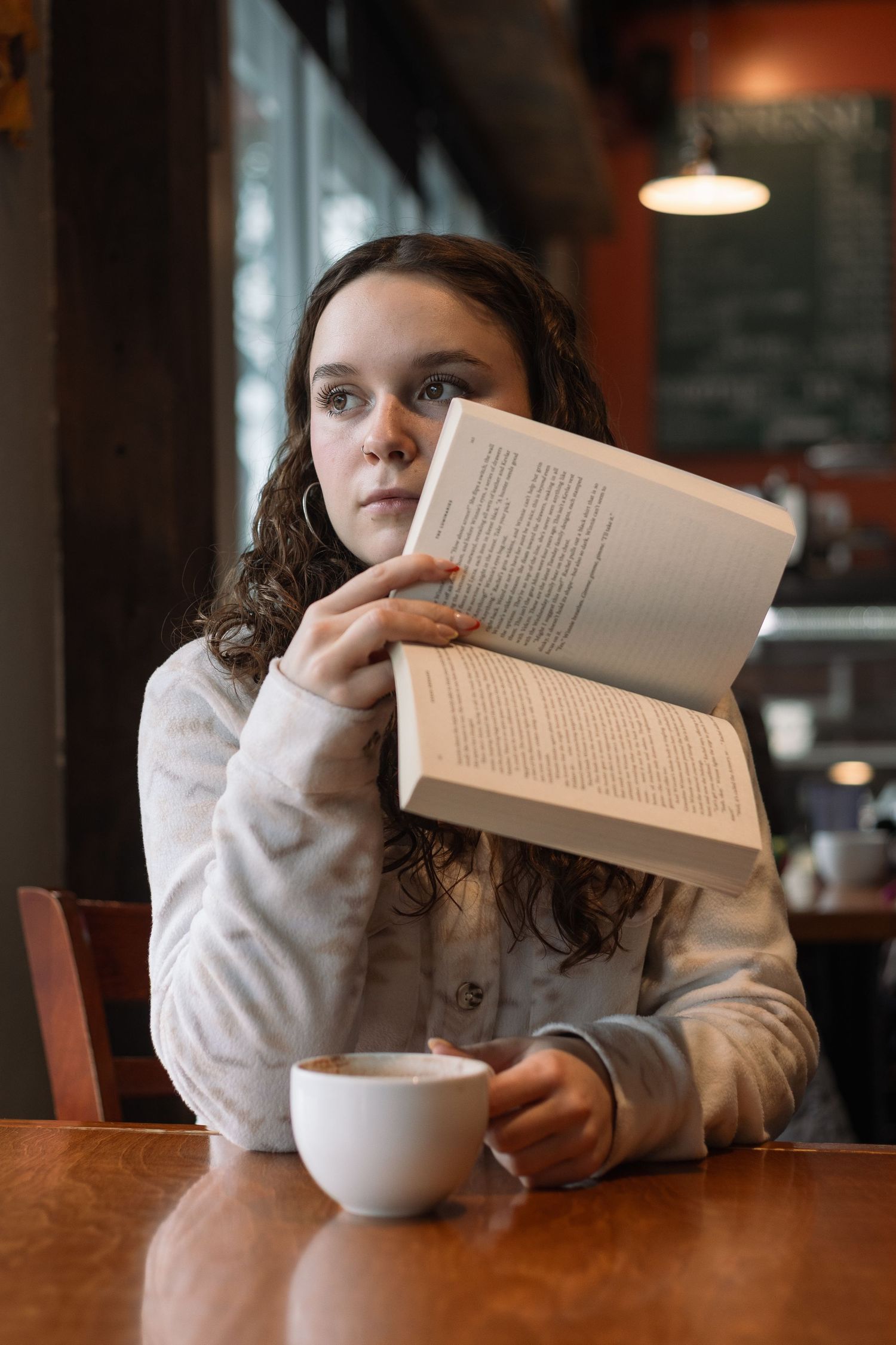 A reader enjoys a cozy moment at a cafe table with a book and white coffee mug.