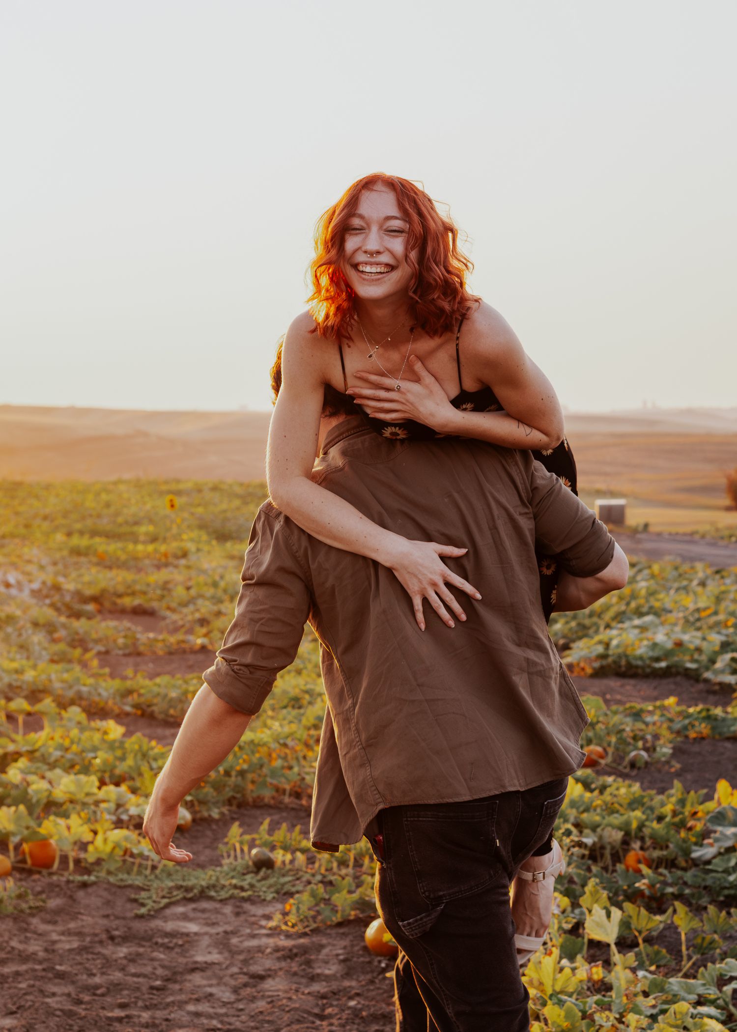 Woman in brown dress laughs joyfully while exploring a rustic pumpkin patch at sunset.