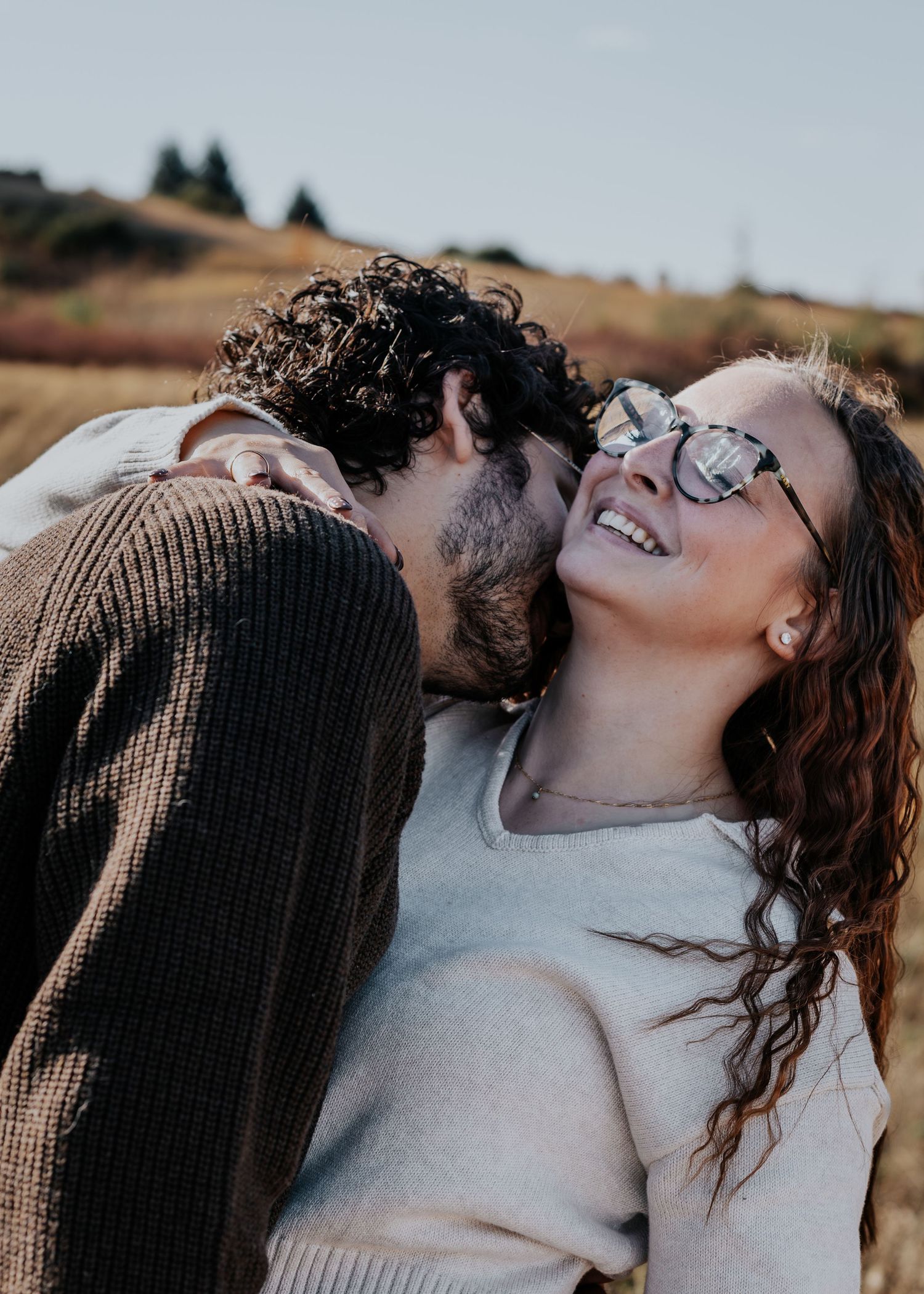 A couple sharing an intimate moment outdoors with one person wearing a brown sweater and the other laughing joyfully.