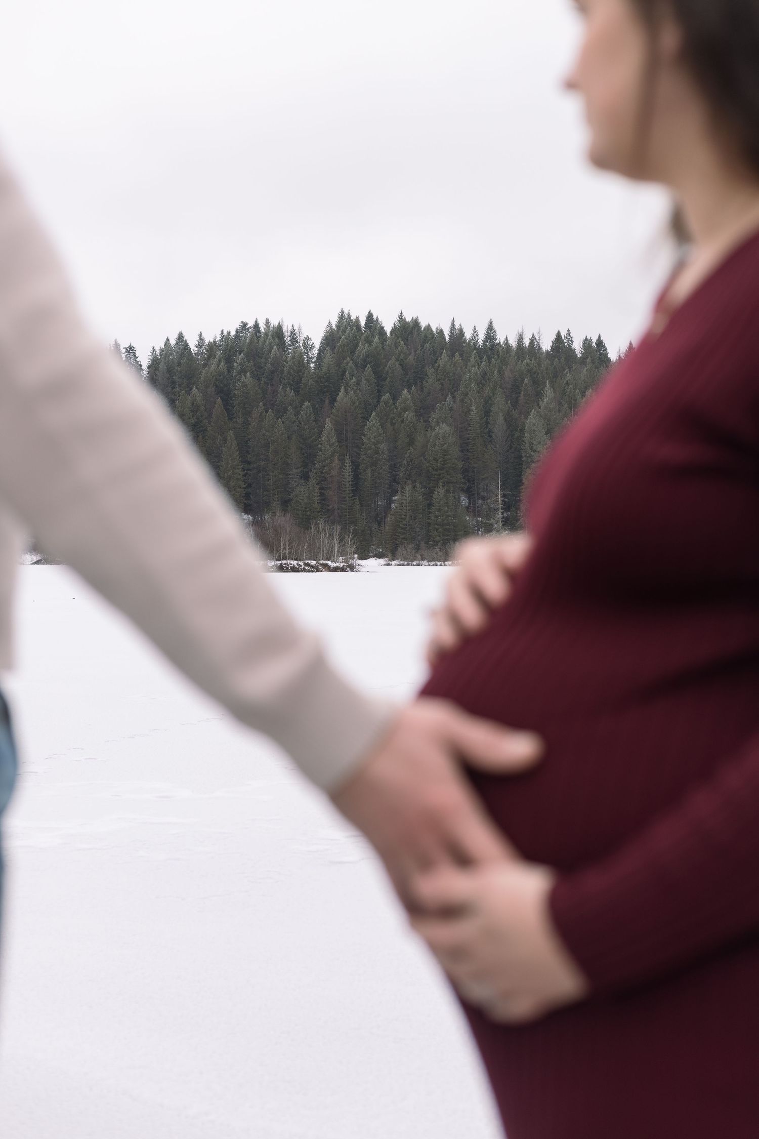 Winter maternity photoshoot by a snow-covered lake with pine forest in background.