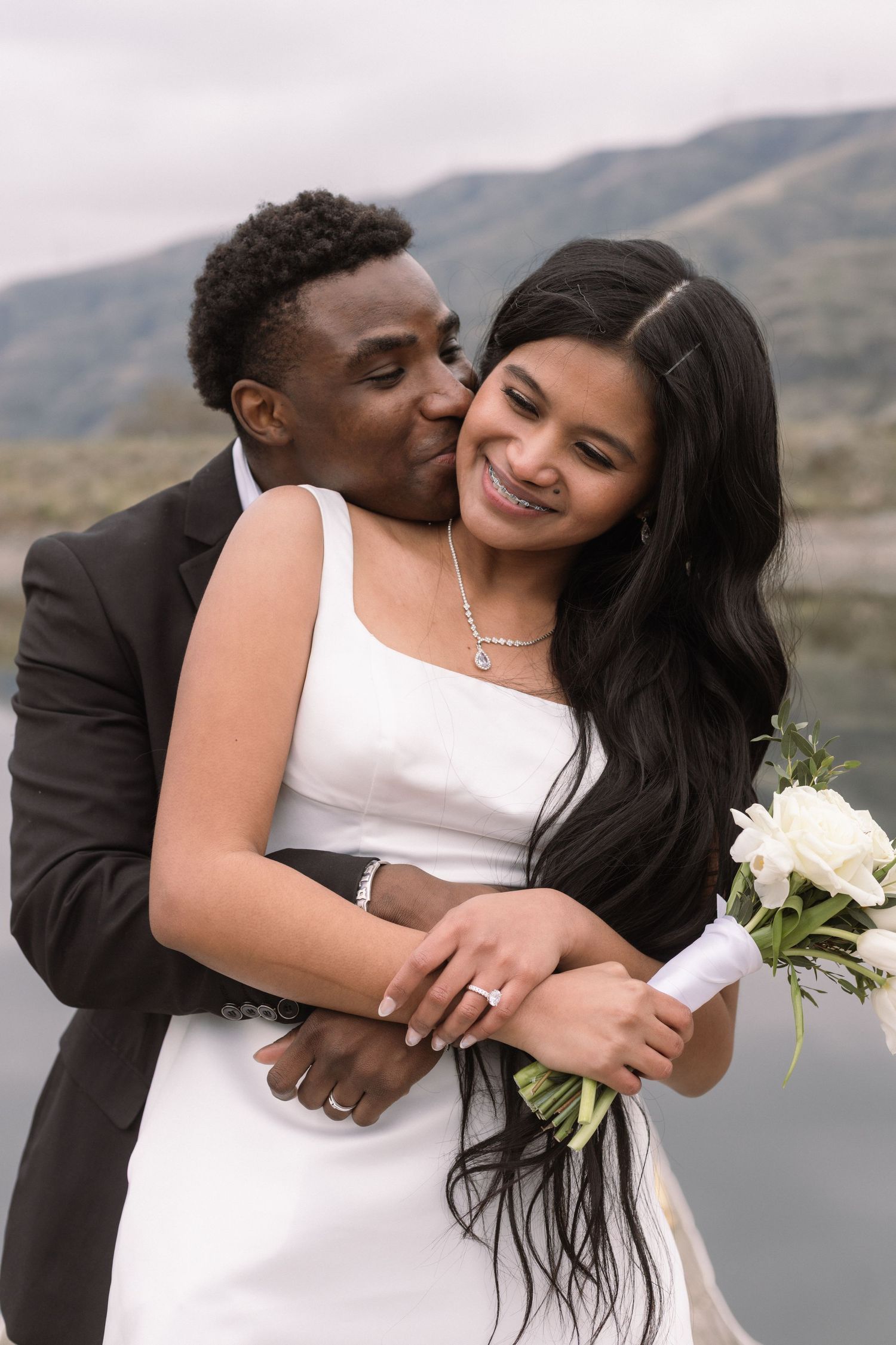 A tender embrace between newlyweds with white flowers against mountains.