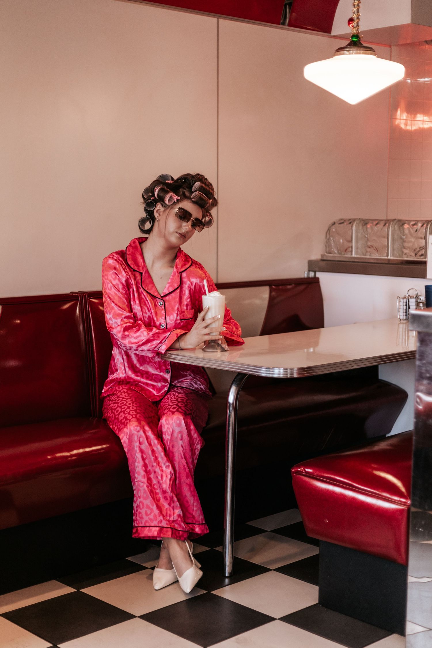 Someone in a shiny pink satin outfit sits at a retro diner booth with black and white checkered floors.