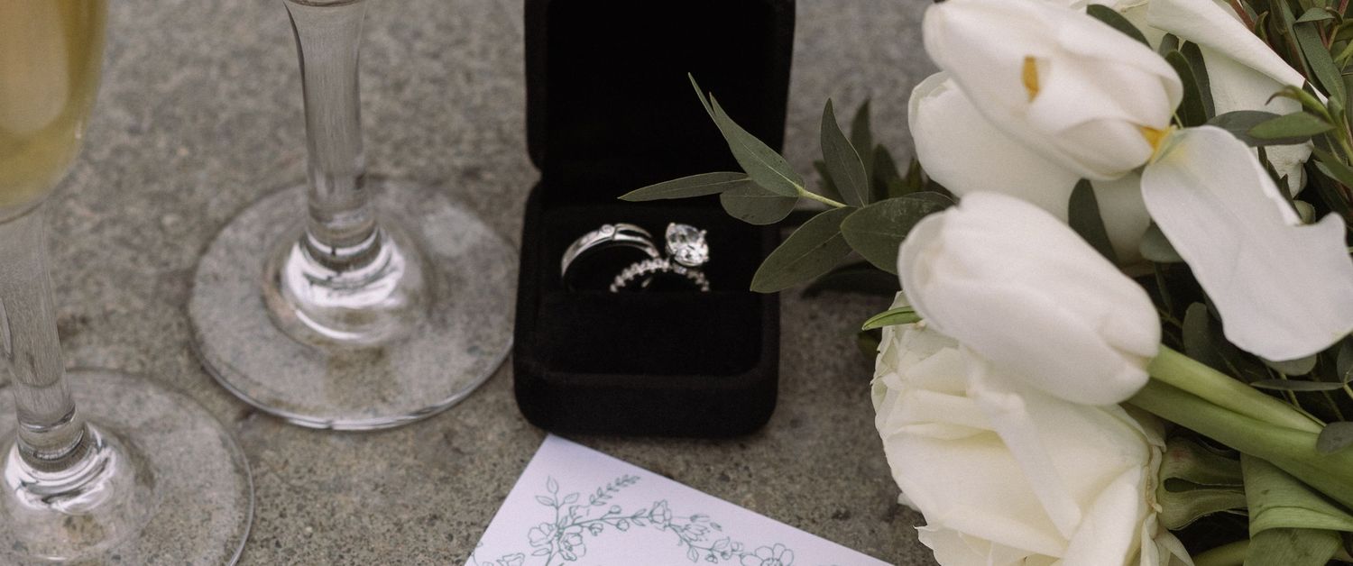Diamond engagement ring in black box beside white tulips and champagne glasses on marble surface.