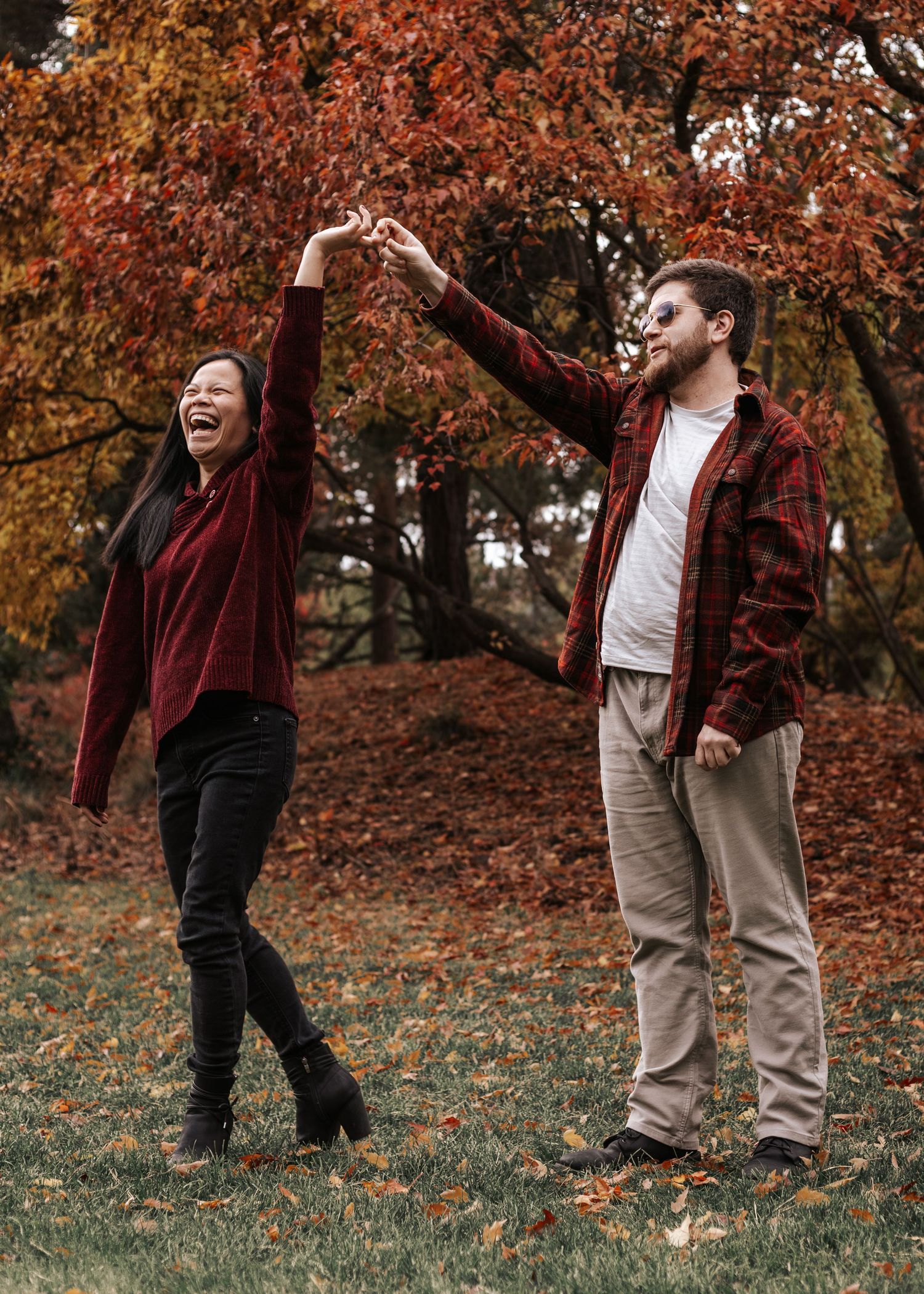 A couple dancing and twirling together in a park surrounded by autumn trees with vibrant red and orange foliage.