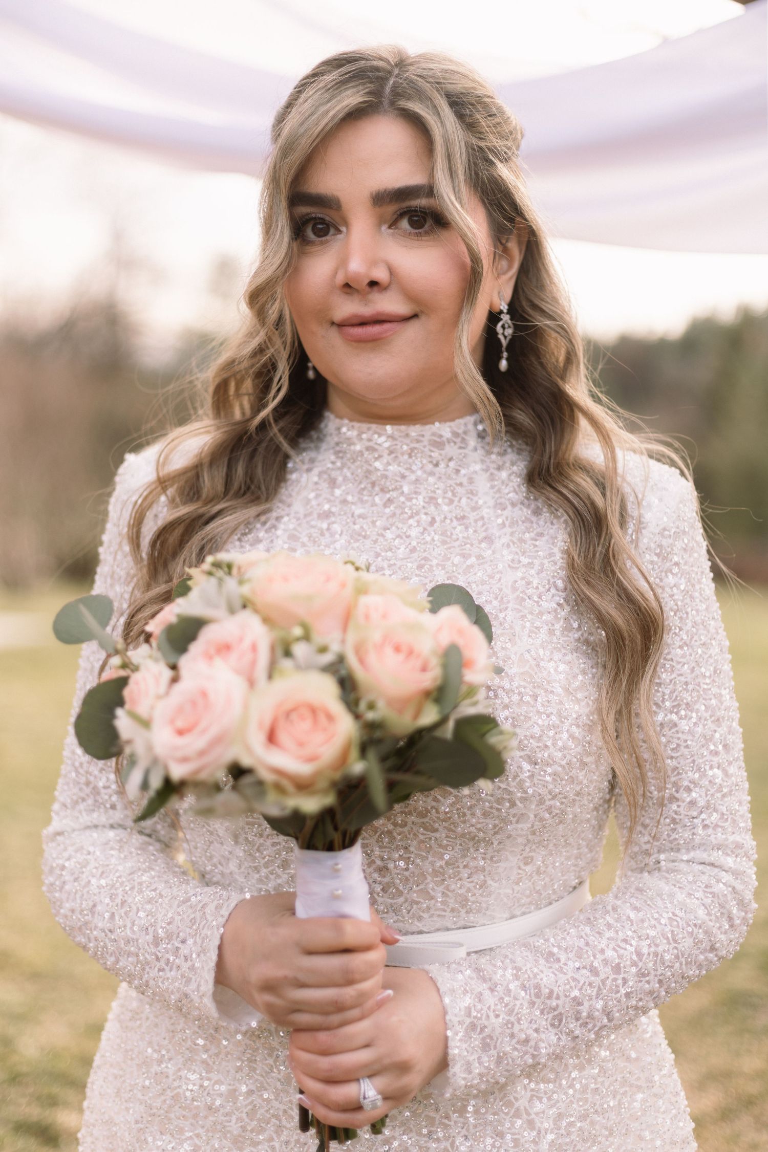 A bride in a long-sleeved sequined wedding dress holds a soft pink and white rose bouquet in an outdoor setting.