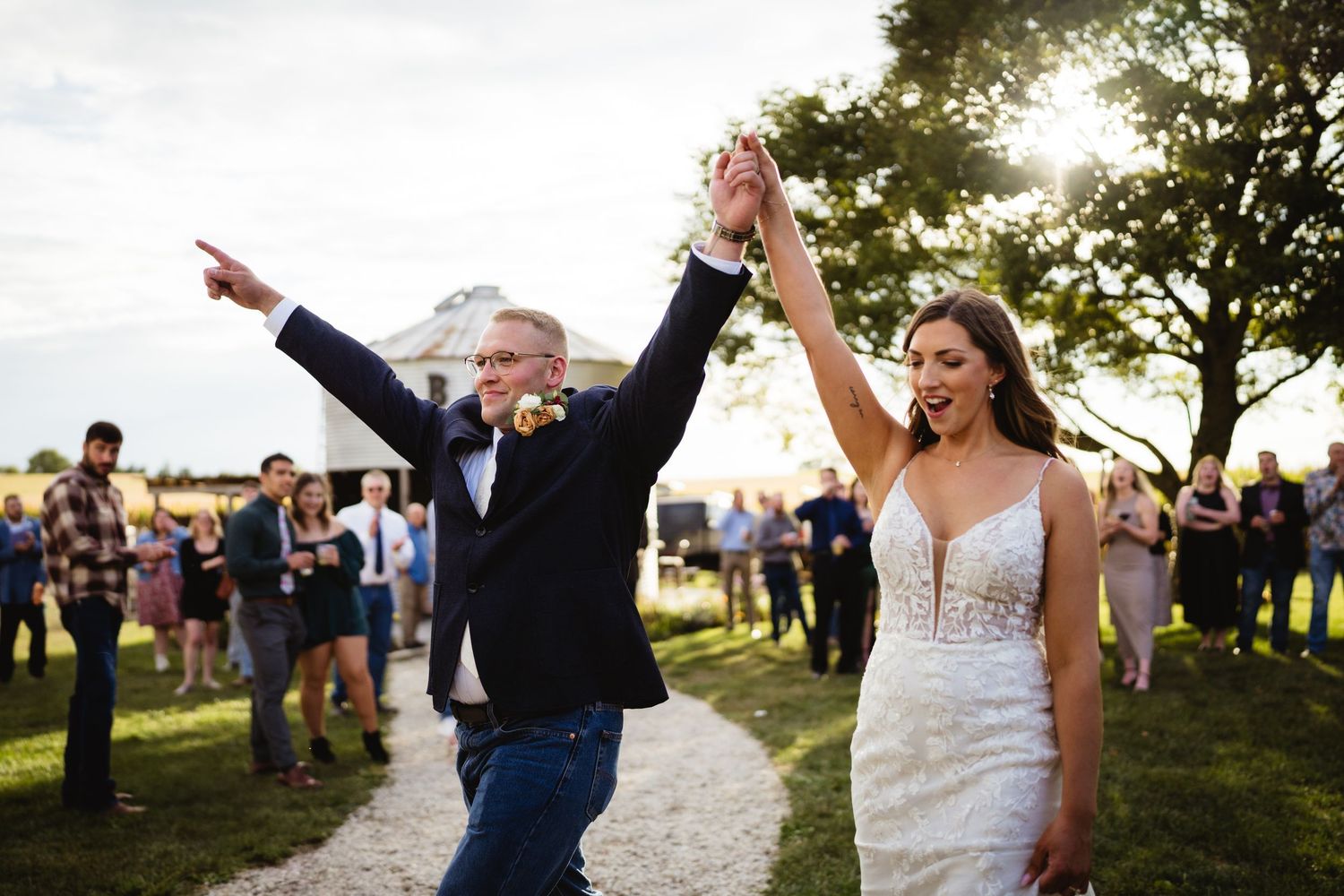 Joyful newlyweds celebrate their wedding reception outdoors, bride in lace gown and groom in navy suit, guests gathered behind.