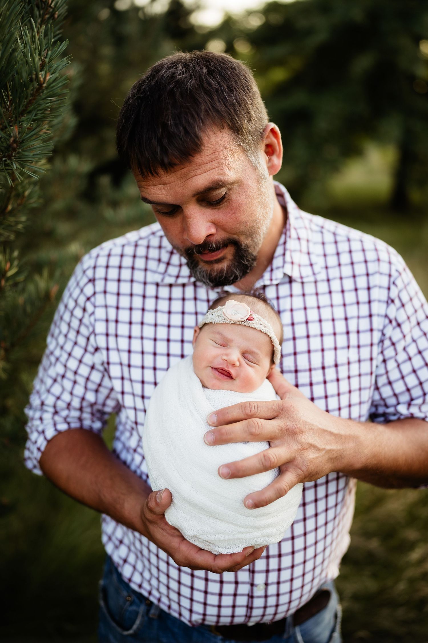 A bearded father tenderly holds his swaddled newborn baby girl outdoors near pine trees.