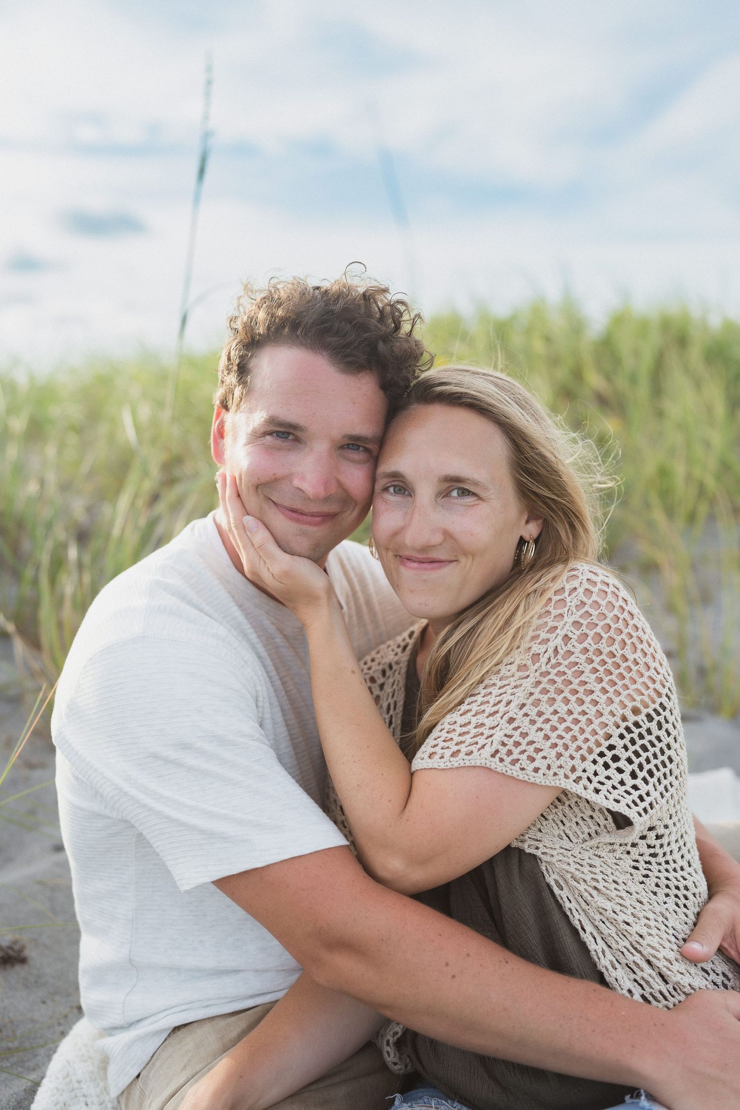 A smiling couple sits together on a sandy beach, with the woman resting her head on the man's shoulder near sea grass.