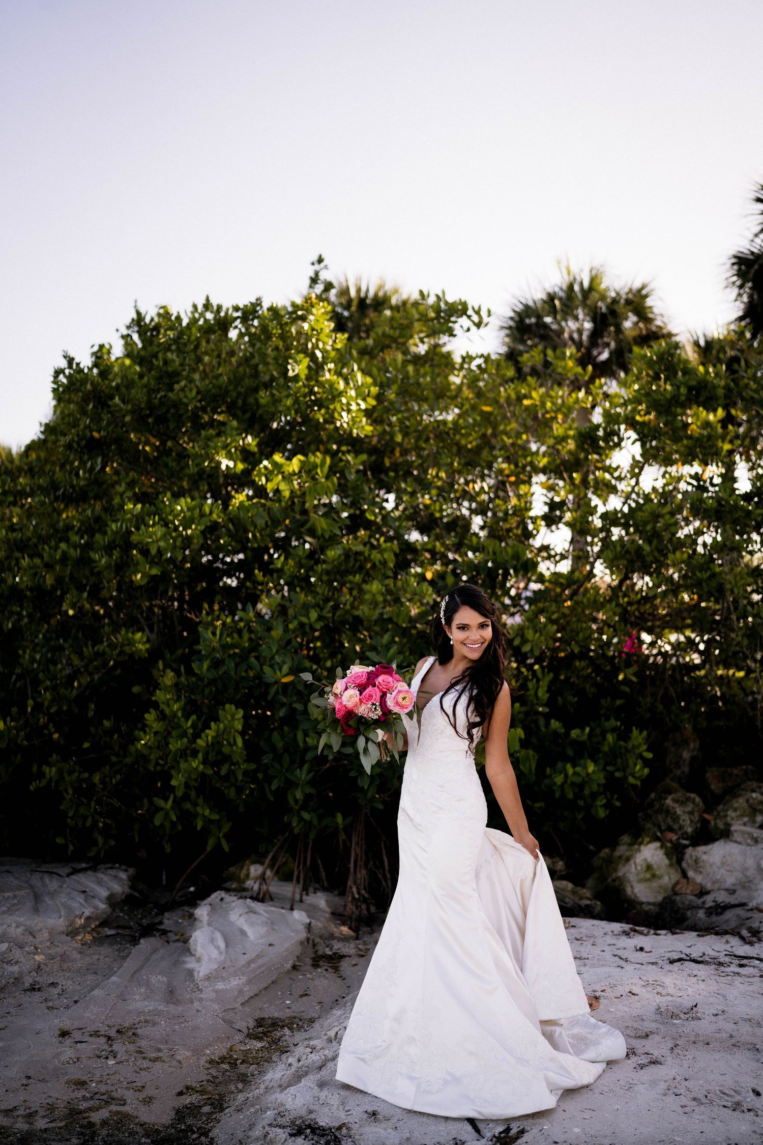 Bride in white mermaid gown holding pink bouquet poses on rocky beach with lush green trees behind her. Hobe Sound Wedding Photographer