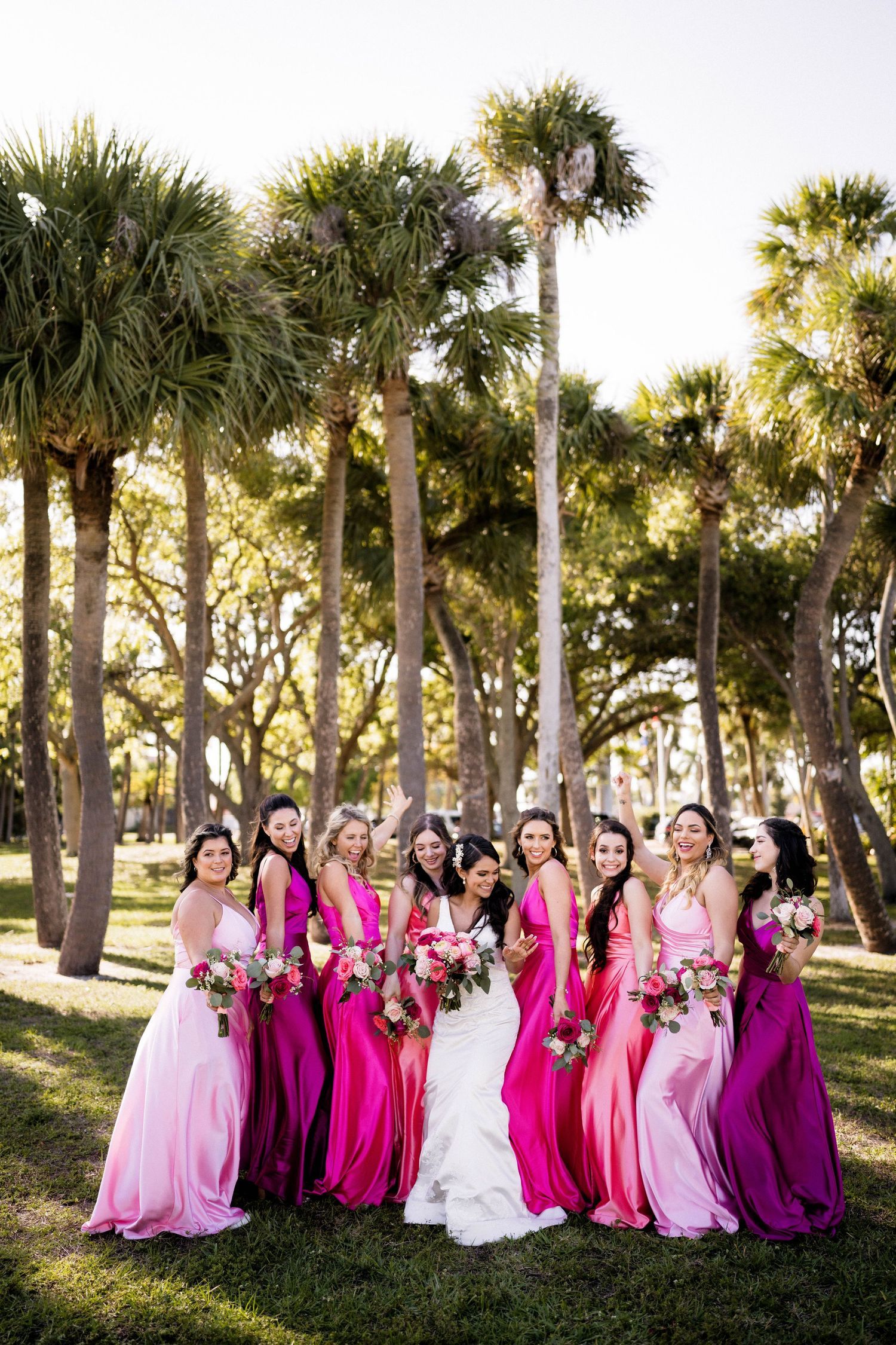 Bride in white gown surrounded by bridesmaids in pink and magenta dresses holding bouquets among palm trees. West Palm Wedding Photographer