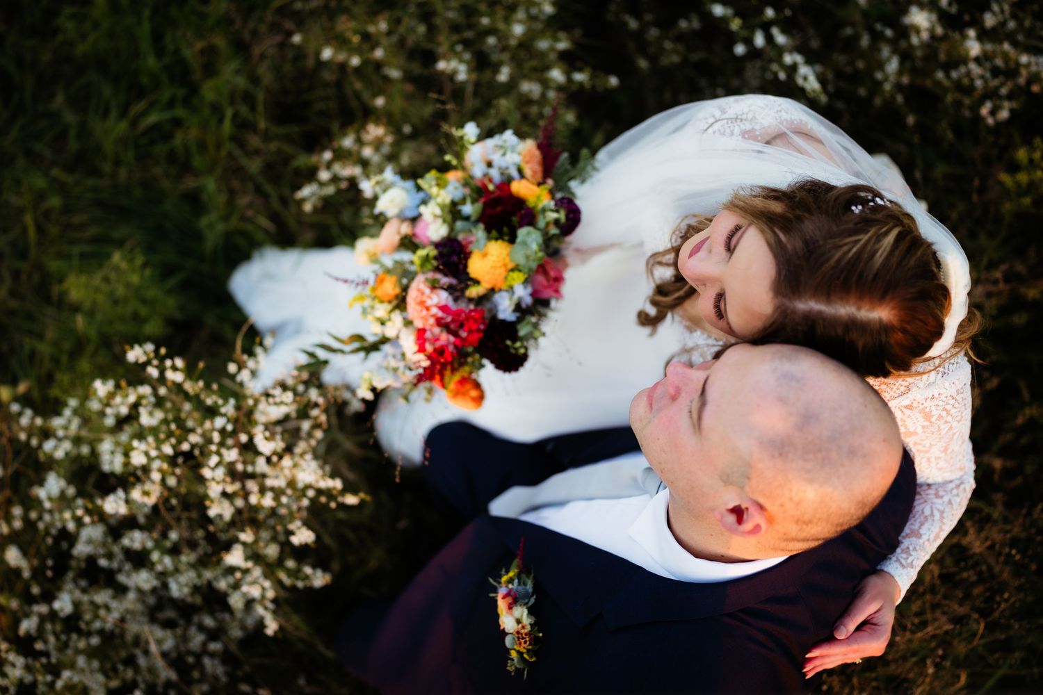 Bride and groom lying in wildflowers, bride resting head on groom's chest holding colorful wedding bouquet.