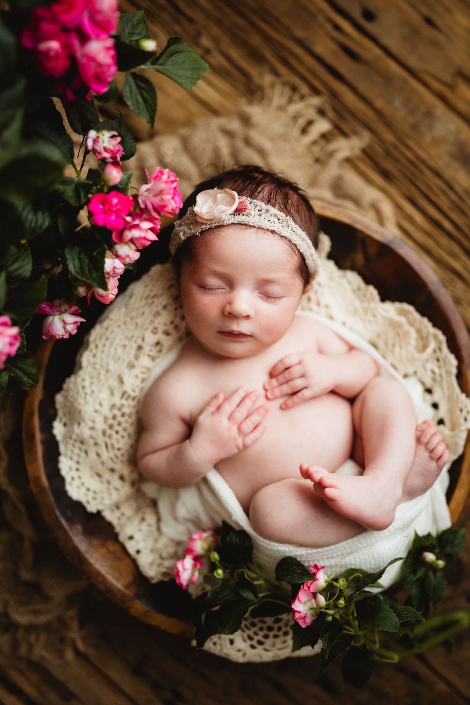 Newborn baby girl sleeping peacefully in a wooden bowl with pink roses and a floral headband on a rustic wood background.