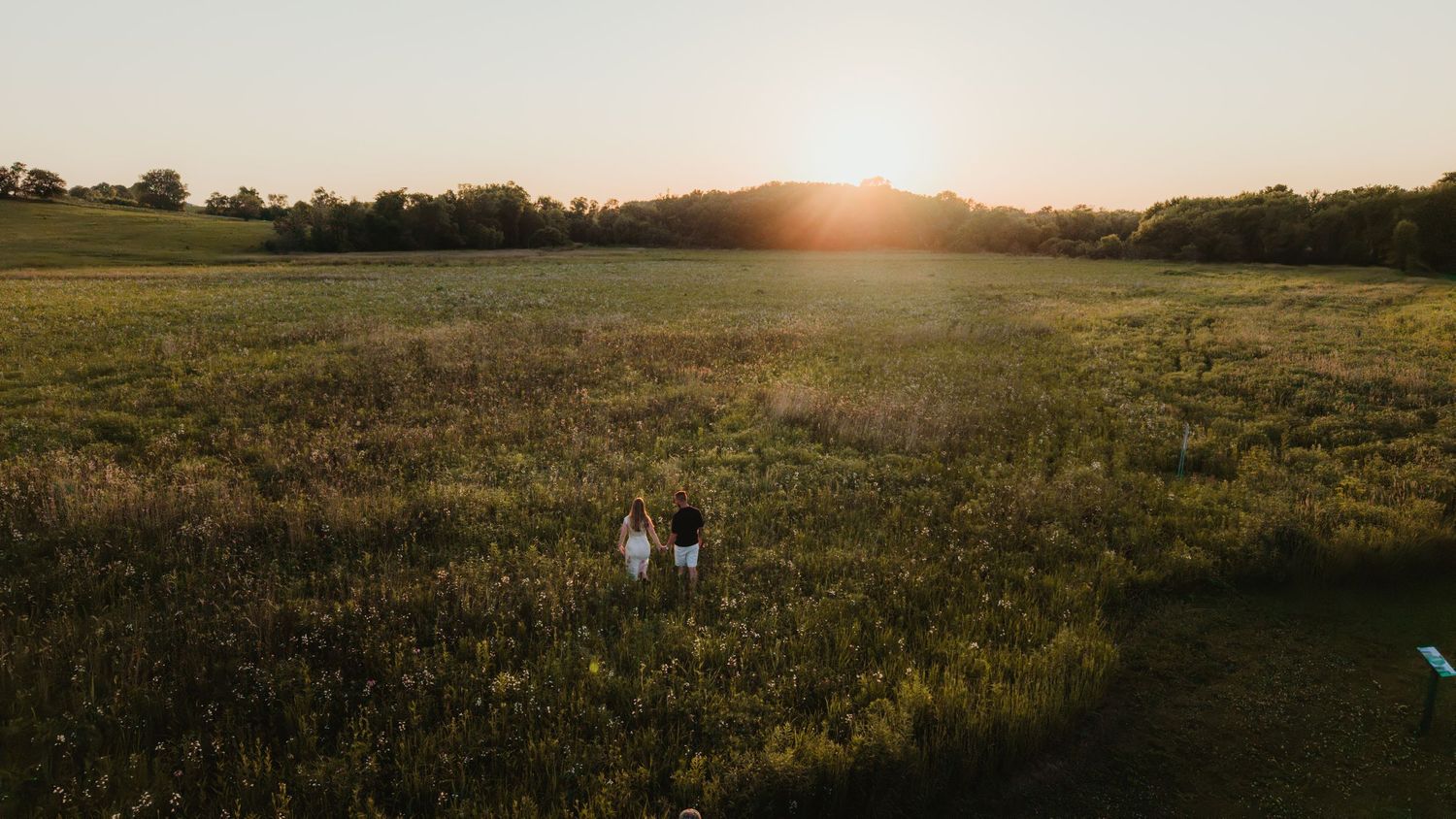 Two figures stand together in a vast grassy meadow at sunset with golden light streaming across the landscape.