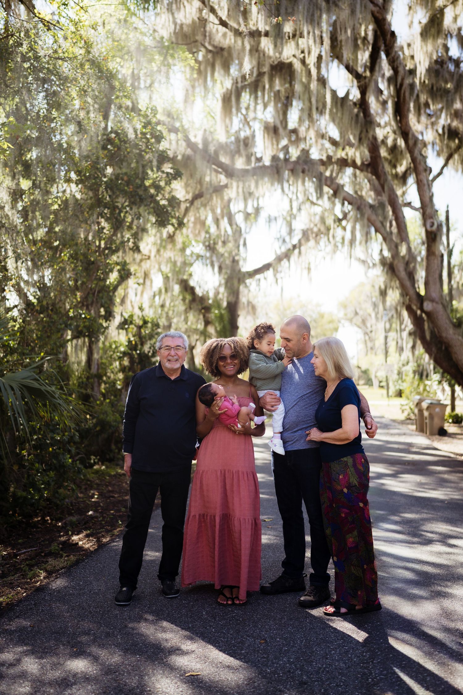 Multigenerational family poses together on a sunlit path under Spanish moss-draped oak trees, with parents holding young children.