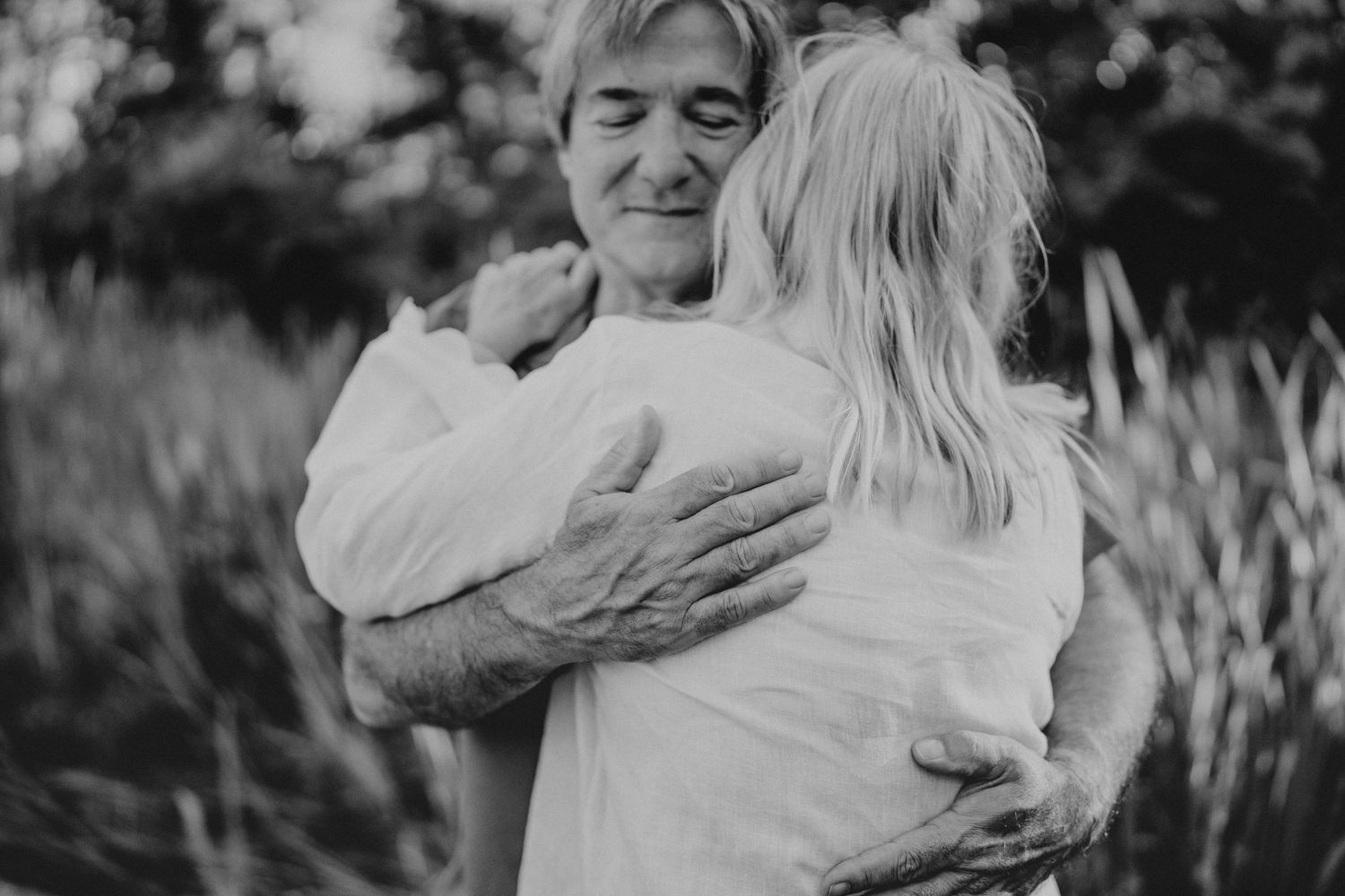 Black and white photo of an elderly man warmly hugging a child outdoors near tall grass.