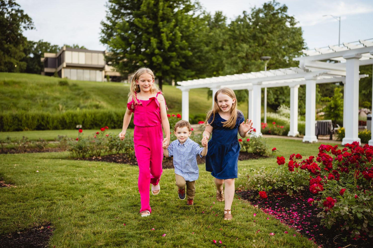 Three happy children running through a lush garden with red roses and white pergola on a sunny day.