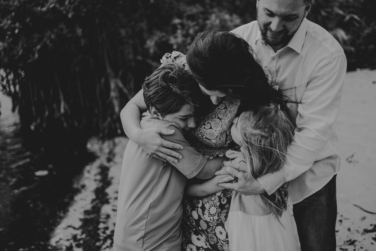 Black and white family photo of parents and children sharing a warm embrace outdoors near lush greenery.