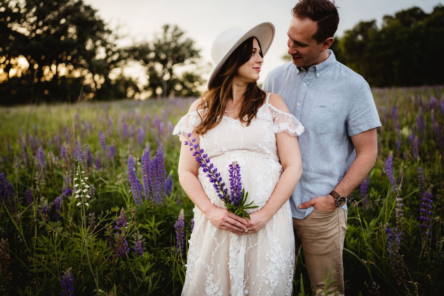 Pregnant woman in white lace dress and hat holds purple lupines with partner in sunny flower field.