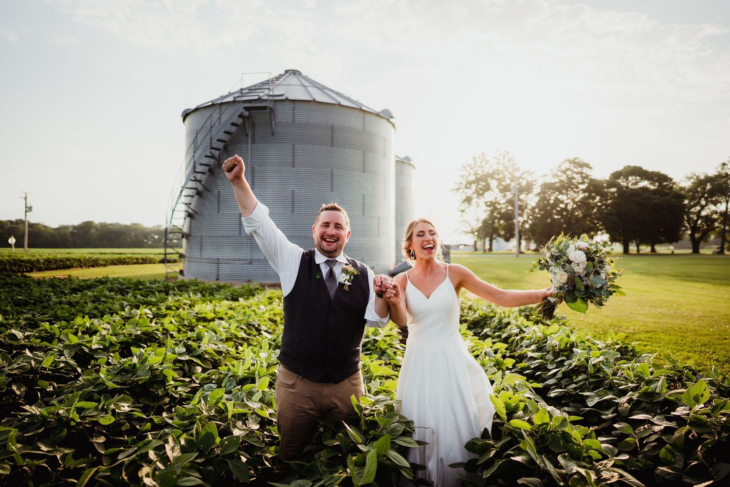 Joyful newlyweds celebrate in a lush green farm field with a grain silo backdrop on their wedding day.