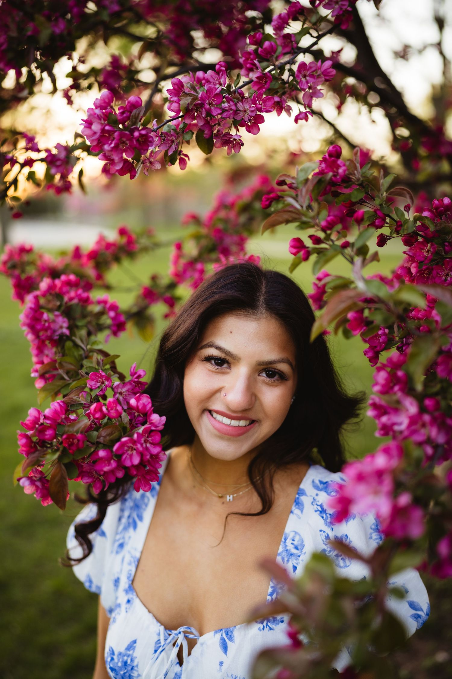 Smiling young woman framed by vibrant pink cherry blossoms in a white floral dress during golden hour.