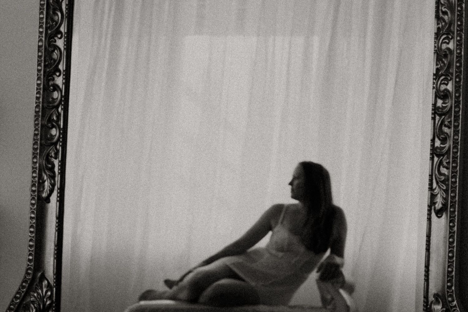 Artistic black and white silhouette photograph framed by elegant ornate borders shows a figure reclining near sheer curtains.