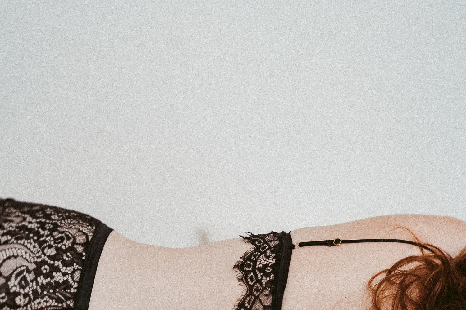 Black lace lingerie detail on pale skin against a light gray background, with auburn hair visible at the edge.