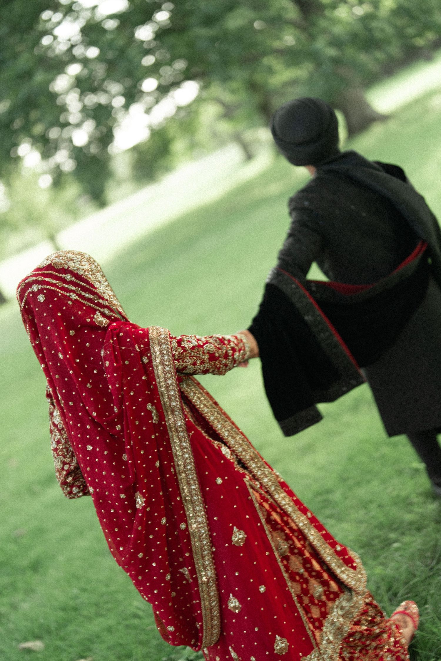 Indian bride in red bridal lehenga holding hands with groom in black sherwani walking through green park.