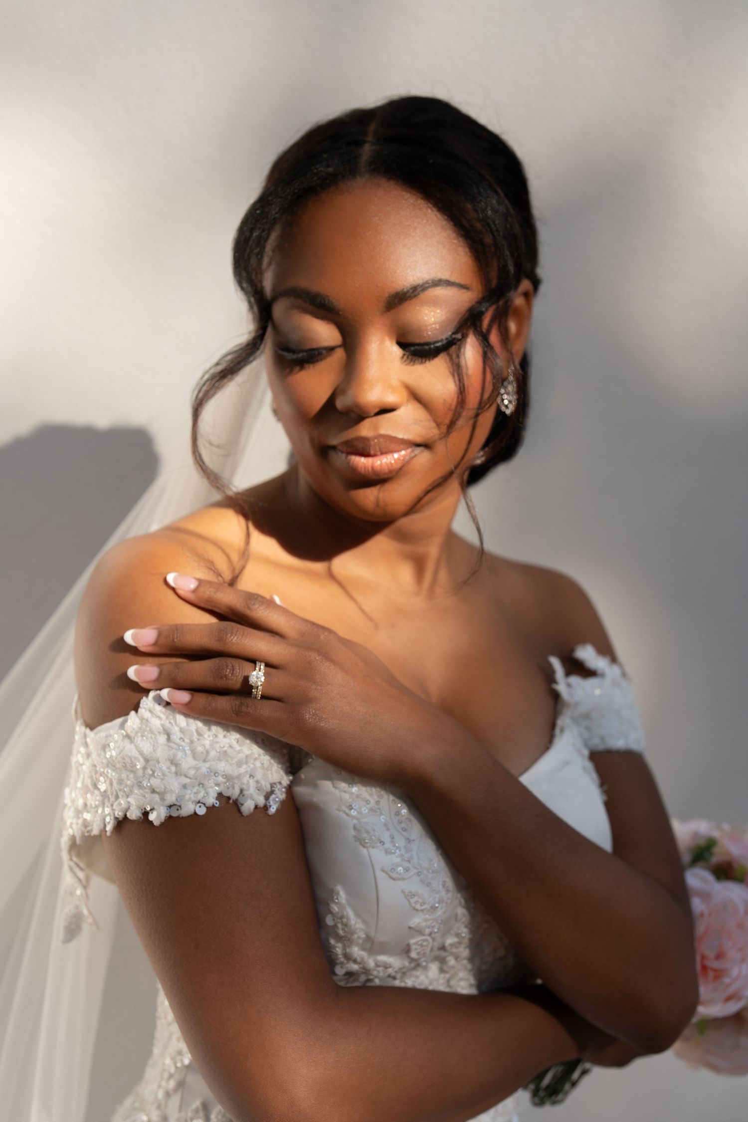 Elegant Black bride in off-shoulder lace wedding gown with veil, wearing diamond earrings, eyes closed, holding bouquet.